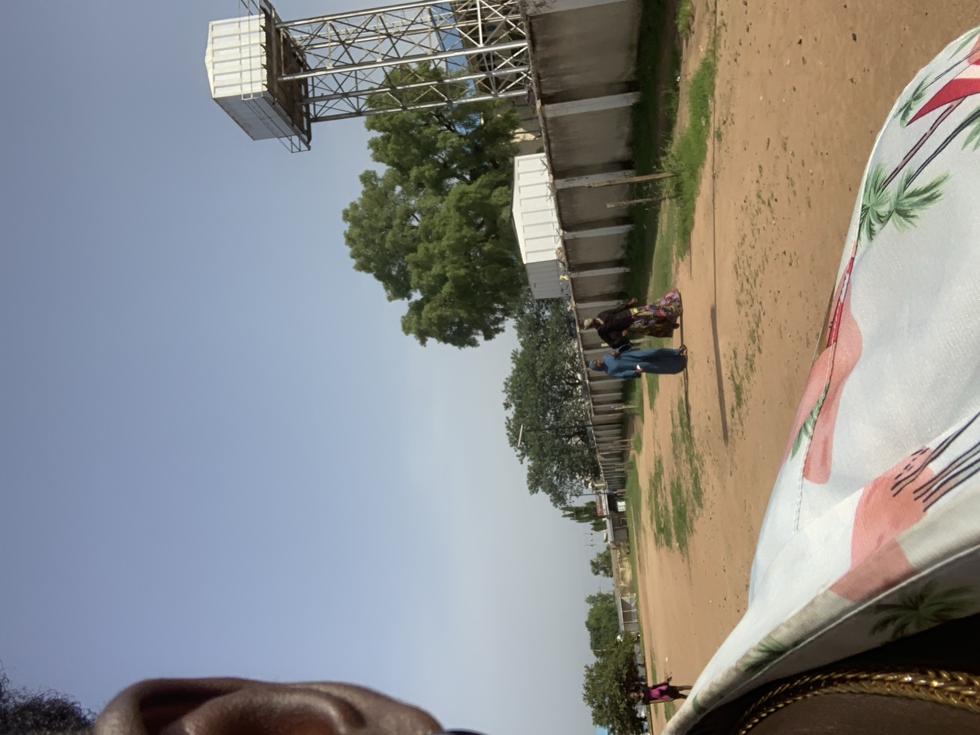 In Jalingo, Nigeria, on a dirt road lined with a concrete wall, two women walk from right to left. The woman on the left wears a blue hijab and dress, while the woman on the right wears a patterned skirt and black top. A young girl in a pink top and dark skirt walks further left, away from the other two women. In the background, a large water tower stands tall against a clear sky. Trees and buildings are visible behind the wall. The foreground is dominated by a person's ear and shoulder, with a colorful garment draped over it.