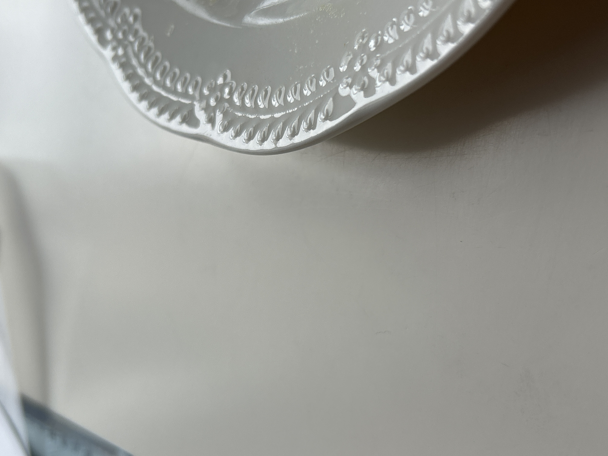 A partial, top-down view of a white, possibly ceramic, plate occupies the upper portion of the frame. The plate features a decorative, beaded edge. The background is a light gray/white surface. The lower portion of the frame is mostly empty space with a hint of white material on the left side and a dark blue/gray rectangular shape below. The location is not identifiable in this image.