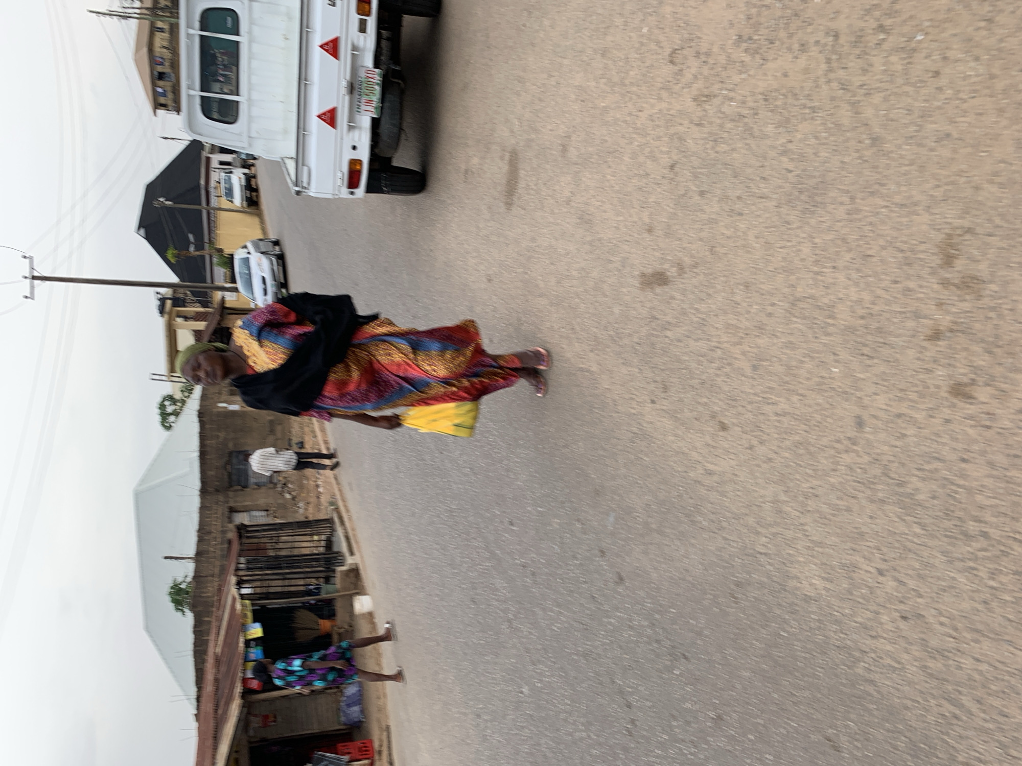 The image shows a street scene in Jos, Nigeria. An elderly woman wearing a colorful patterned dress and a black shawl is walking along the road. She carries a yellow bag. A white truck is parked on the side of the road. Other people are visible in the background, along with roadside businesses and buildings. Utility poles line the street. The weather appears overcast.