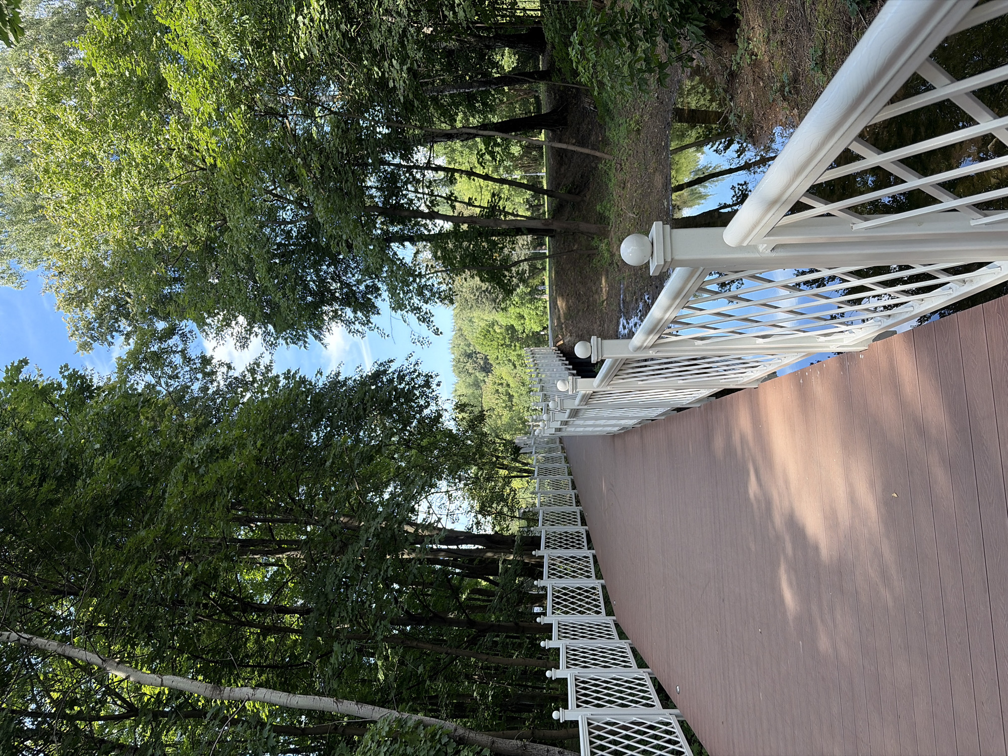 This is a full-shot, eye-level, outdoor, vertical photograph of a wooden boardwalk with white railings that curves to the left through a park. The boardwalk is made of brown planks and is lined on both sides by a decorative white lattice railing with spherical finials. Sunlight dapples the surface of the boardwalk, casting shadows from unseen trees. On the right side of the boardwalk, a shallow, muddy stream reflects the surrounding trees and sky. The park itself is densely wooded with mature trees, their green leaves filling the upper portion of the frame, contrasting with a bright blue sky with scattered white clouds. In the distance, the terrain slopes upward, covered with more trees. The scene appears to be of a walking path designed to traverse a natural landscape within a city park. This boardwalk is located in the Arkhangelskoye Estate Museum-Reserve in Krasnogorsk, Moscow Oblast, Russia. There are no people or animals visible in the image.