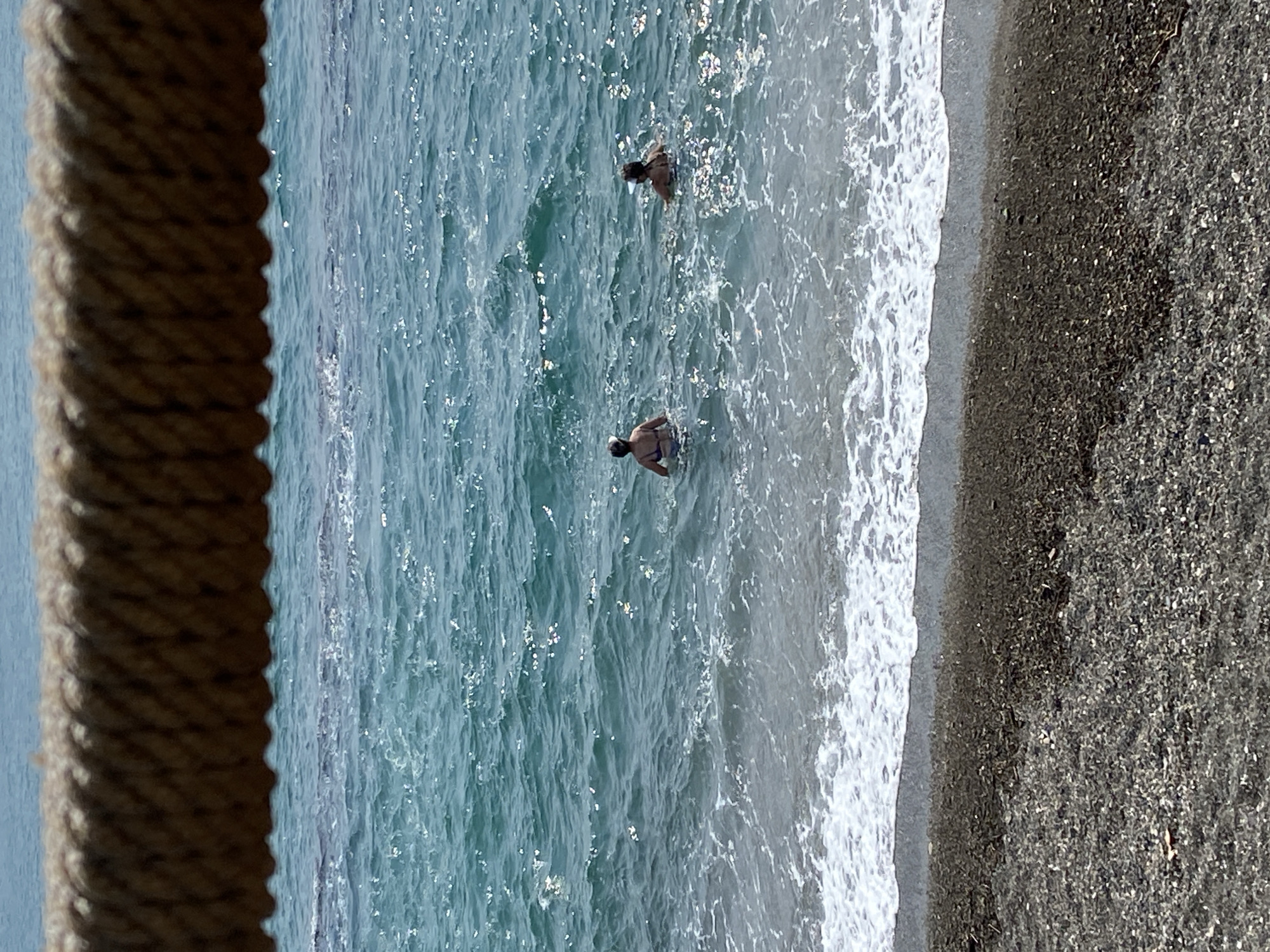 The image depicts a beach scene in Chabkwara, Abkhazia. In the foreground, a thick, woven rope stretches horizontally across the frame, obscuring the view of the upper part of the sea. The sea itself is a vibrant turquoise, with small waves breaking near the shore, creating white foam. The beach is composed of dark grey pebbles and sand. Two individuals are visible in the water, both appearing to be women wearing swimsuits. One is closer to the left edge of the frame, facing away from the viewer, and the other is further to the right, also facing away. Both are wading in the sea, with the water reaching approximately chest height for the person on the left and waist height for the person on the right. The sunlight reflects off the water's surface, creating a shimmering effect.