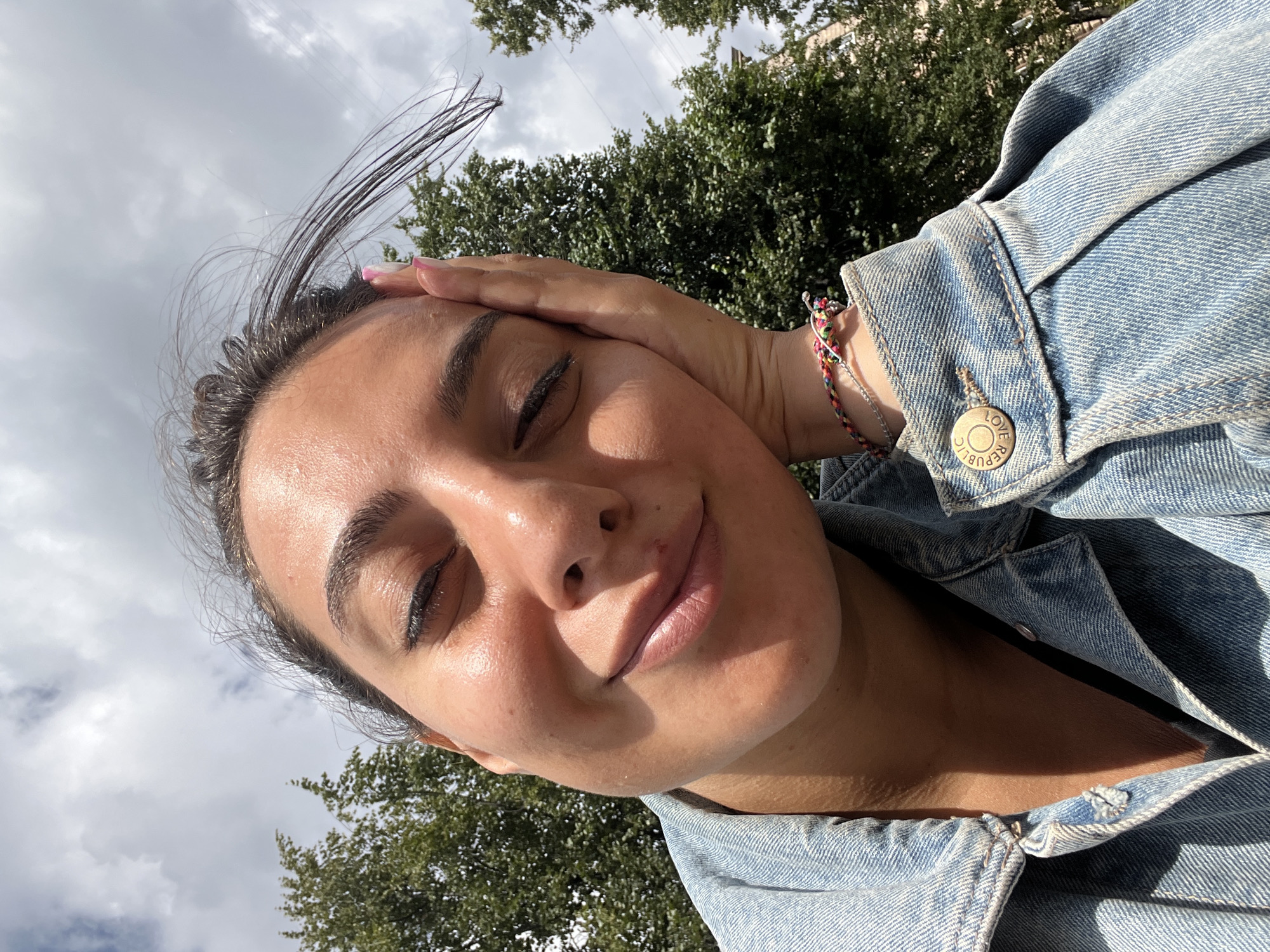 A young woman is taking a selfie outdoors. She has dark hair and is wearing a denim jacket. Her left hand is resting on her cheek, and she is smiling with her eyes closed. The background consists of trees and a cloudy sky. The image appears to have been taken in an urban park or street setting in Saint Petersburg, Russia, based on the style of the trees and the general atmosphere.