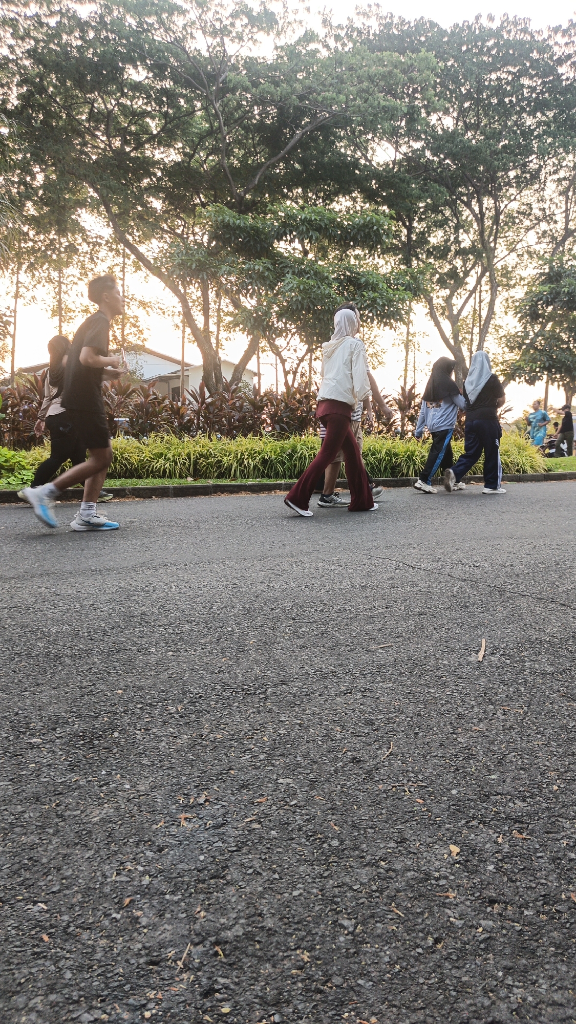In Bacin, Indonesia, a group of people are walking and jogging on a paved path lined with trees and decorative plants. The sun is setting, casting a warm glow on the scene. In the foreground, a young man in a black t-shirt and shorts is jogging with a determined stride, his light blue and white athletic shoes striking the pavement. Behind him, two other individuals, one in a beige t-shirt and the other in a dark t-shirt, walk together. Further down the path, a person in a white hooded jacket and maroon pants is walking, while two others in hijabs and athletic wear follow closely. The background is filled with lush greenery, with tall trees forming a canopy overhead. The overall atmosphere is one of casual recreation and outdoor activity.