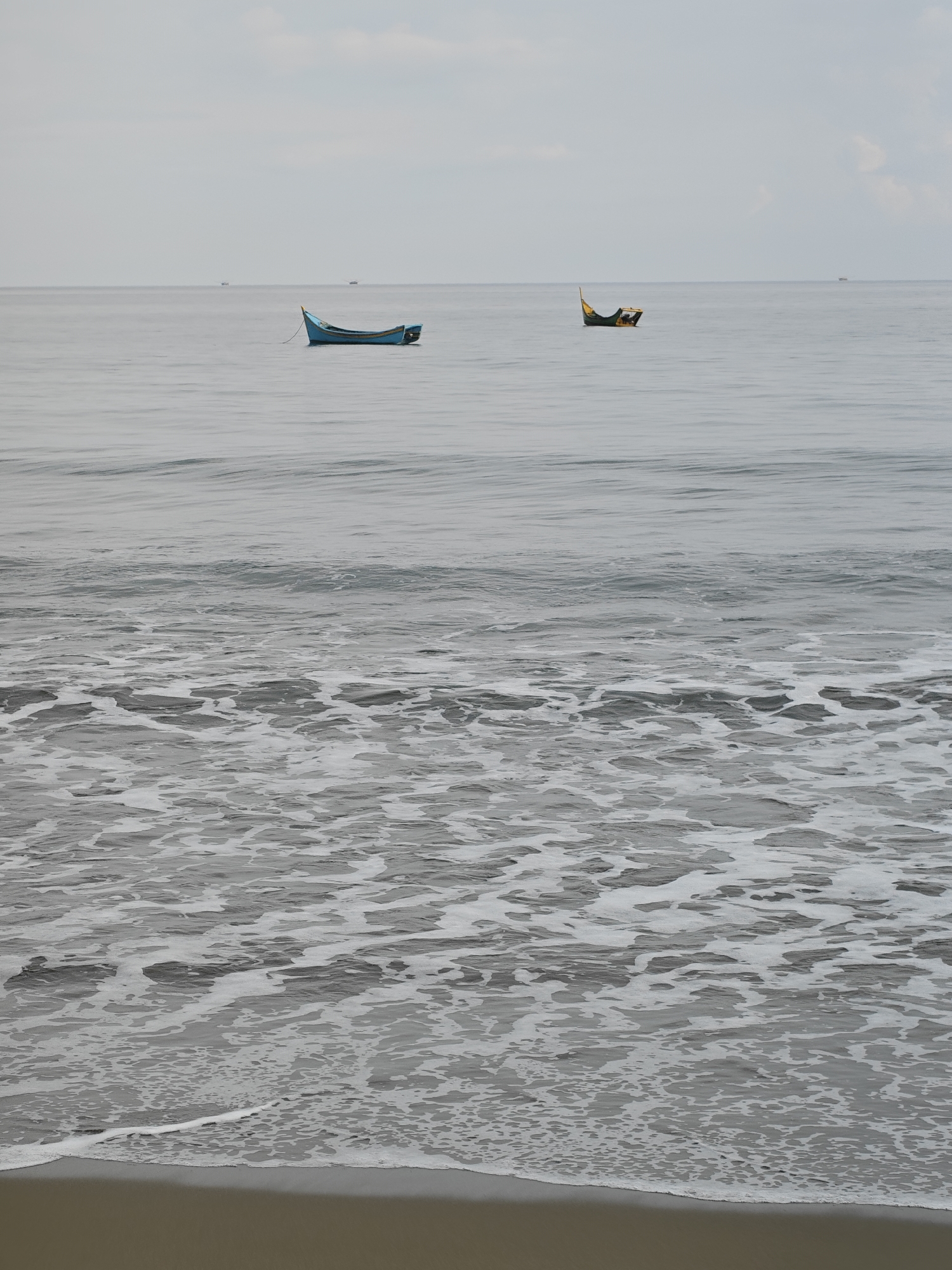 In Curehtunong, Indonesia, two small fishing boats are moored on a calm, grey sea under an overcast sky. The boat on the left is a bright blue, narrow vessel, while the boat on the right is dark green with yellow accents. The water is mostly still, with gentle ripples on the surface. Closer to the shore, the water breaks into white foam as it washes onto a sandy beach. The overall atmosphere is serene and quiet.