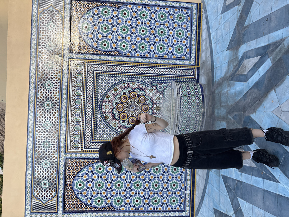 A person stands next to a decorative wall, their body angled towards the right. They wear a white t-shirt, black cargo pants with grommet detailing, and black sneakers. A baseball cap adorns their head, and a necklace with a cross hangs around their neck. Tattoos are visible on their arms. The wall features intricate tile patterns in shades of blue, green, and yellow. The ground has blue and gray geometric designs.