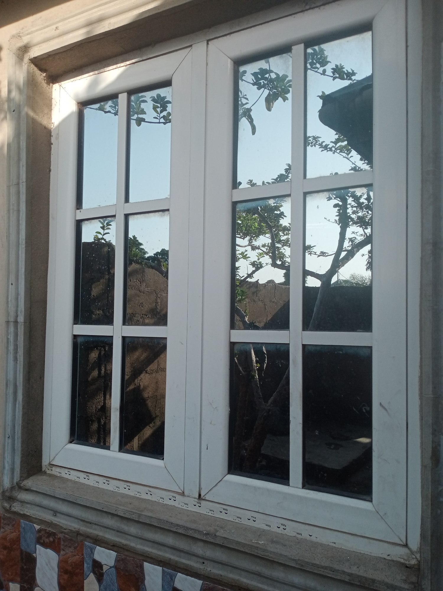 The image shows a close-up of a white-framed window with multiple panes. The glass reflects the sky, trees, and a portion of a dark roof. The window is set into a textured, light-colored wall. Below the window, there is a tiled surface with a pattern of brown, white, and blue tiles. The overall scene suggests an outdoor setting, likely a residential or commercial building in Jalingo, Nigeria, given the context. No people are visible in the image.