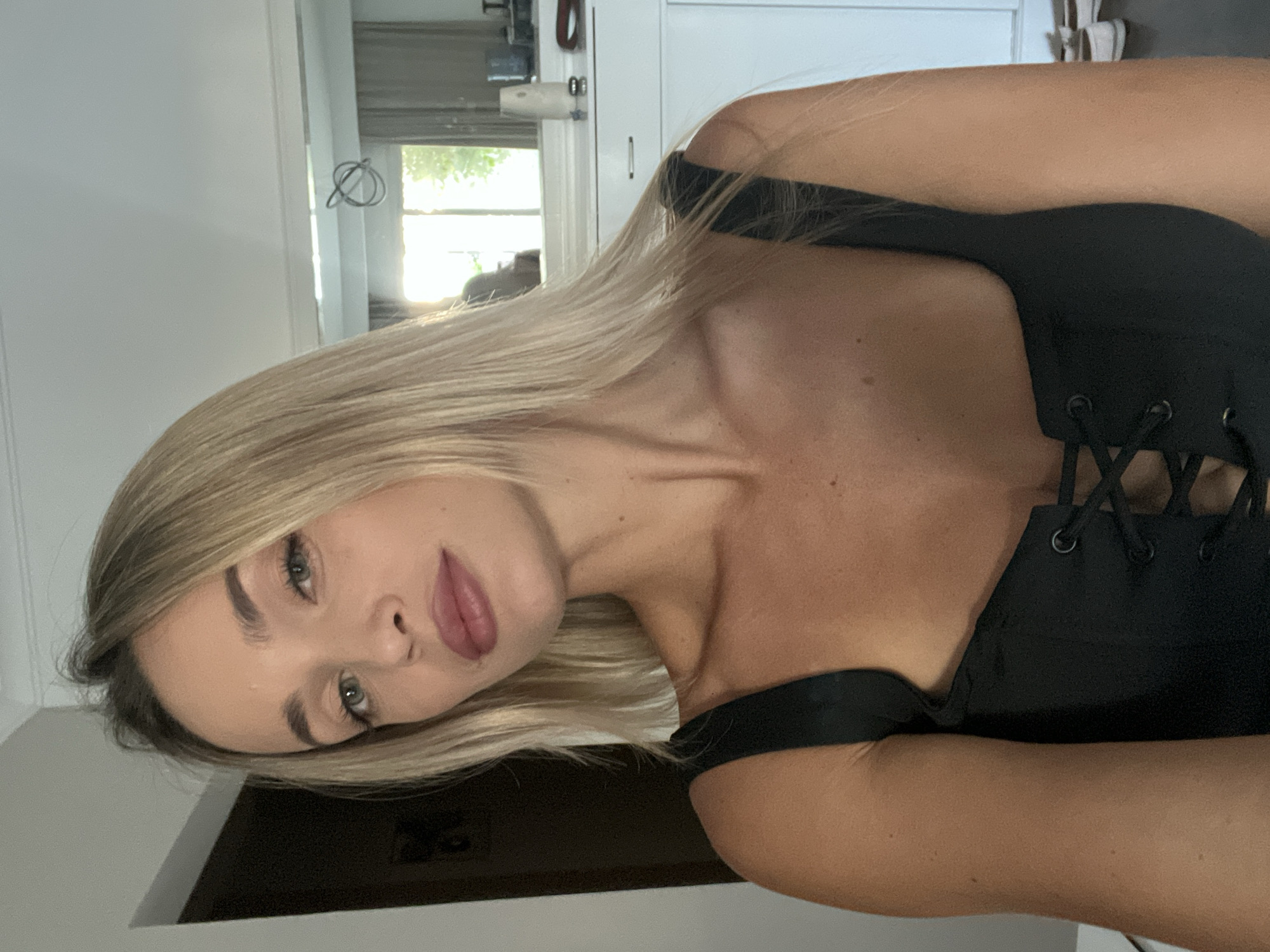 A blonde woman with green eyes and pink lipstick is captured in a selfie. She is wearing a black corset-style top with lace-up detailing. The background suggests an indoor setting, possibly a room with a mirror reflecting a window that allows natural light to enter, indicating it is daytime. The environment appears to be a well-lit room with furniture, including a dresser or cabinet. The overall scene is casual and personal. There is no indication that this image is related to Sharm El Sheikh, Egypt.