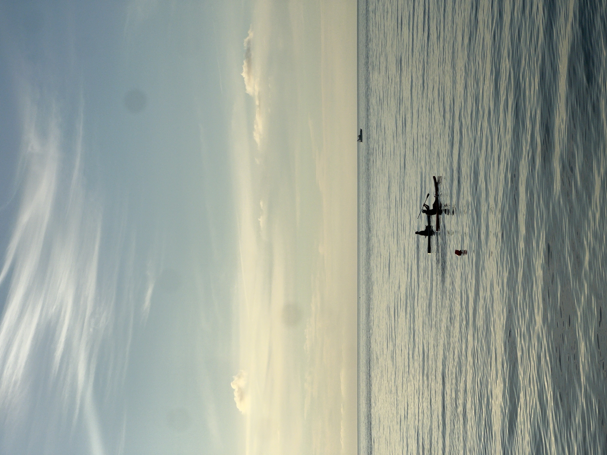 Two individuals are paddleboarding on calm water. One person is in front and is using a paddle. The other person is behind and appears to be sitting or kneeling on the paddleboard. The paddleboarders are silhouetted against the water, and their specific features are not discernible. In the distance, a small boat is visible on the horizon. The sky is partly cloudy with a light blue hue. The overall scene suggests a serene outdoor activity, likely taking place at sea or a large body of water near Batumi, Georgia.