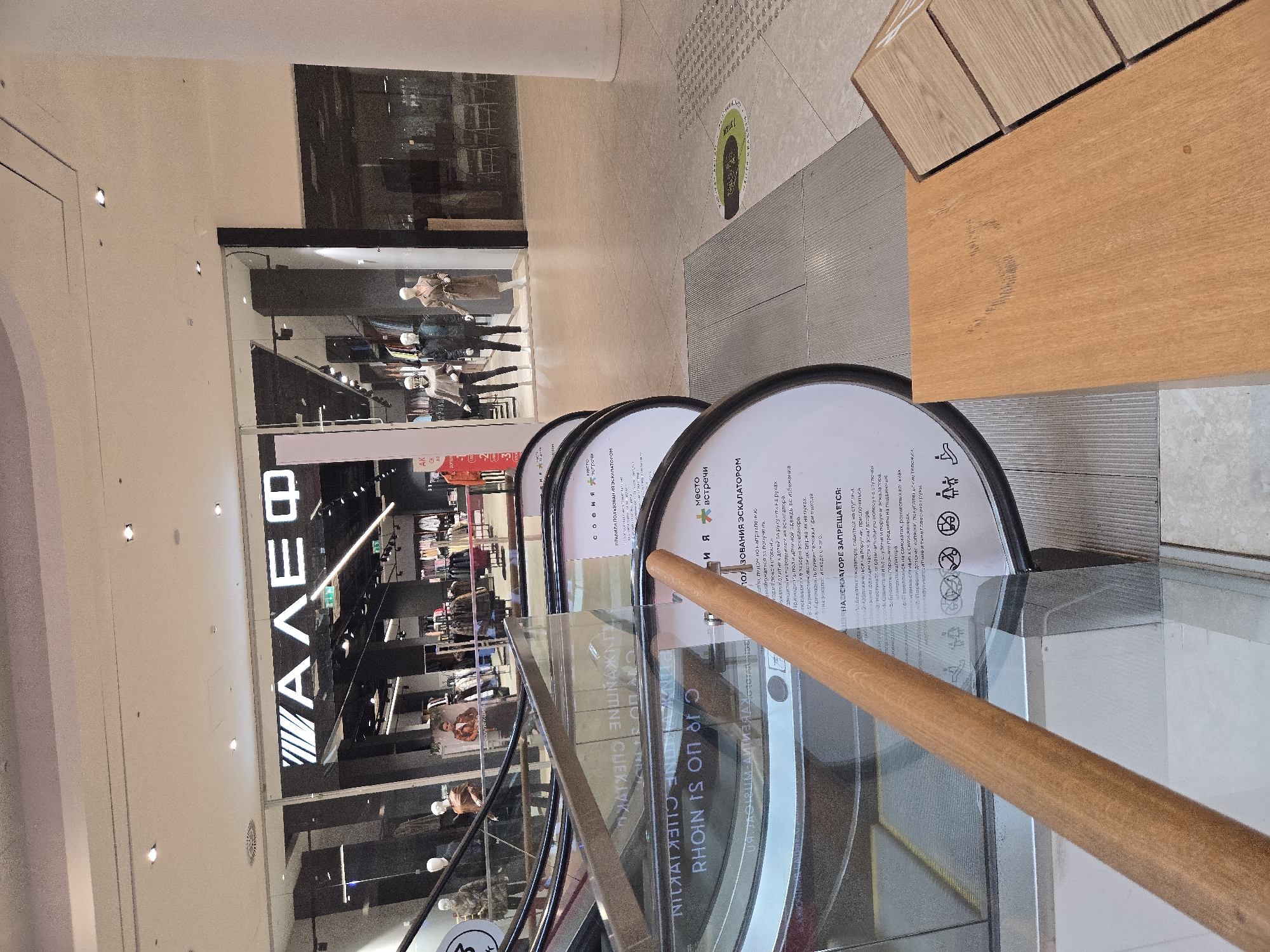 This media file captures a view inside a modern shopping mall, likely in Moscow, Russia. The scene is dominated by the polished floors, glass railings, and the illuminated signage of a retail store. The focal point of the image is an escalator, its rounded edges and glass barriers prominently featured. A light-colored wooden handrail guides the eye along the escalator's path.
Several mannequins are visible in the store window, displaying clothing items, suggesting a commercial environment. The store's signage, prominently displaying the Cyrillic letters "АЛЕФ," indicates its name. Several circular signs with text in Russian are affixed to the escalator's glass barriers. These signs appear to be safety instructions and rules for using the escalator, with headings such as "ПРАВИЛА ПОЛЬЗОВАНИЯ ЭСКАЛАТОРОМ" (Rules for Using the Escalator) and "НА ЭСКАЛАТОРЕ ЗАПРЕЩАЕТСЯ" (It is Prohibited on the Escalator).
The lighting suggests it is daytime, with the bright interior lights of the mall illuminating the space. The overall atmosphere is clean and contemporary, typical of a retail setting. No people are clearly visible in the immediate foreground, but the presence of mannequins and store displays implies a public space with shopper activity. The orientation of the image is somewhat tilted, giving a dynamic perspective of the scene.
