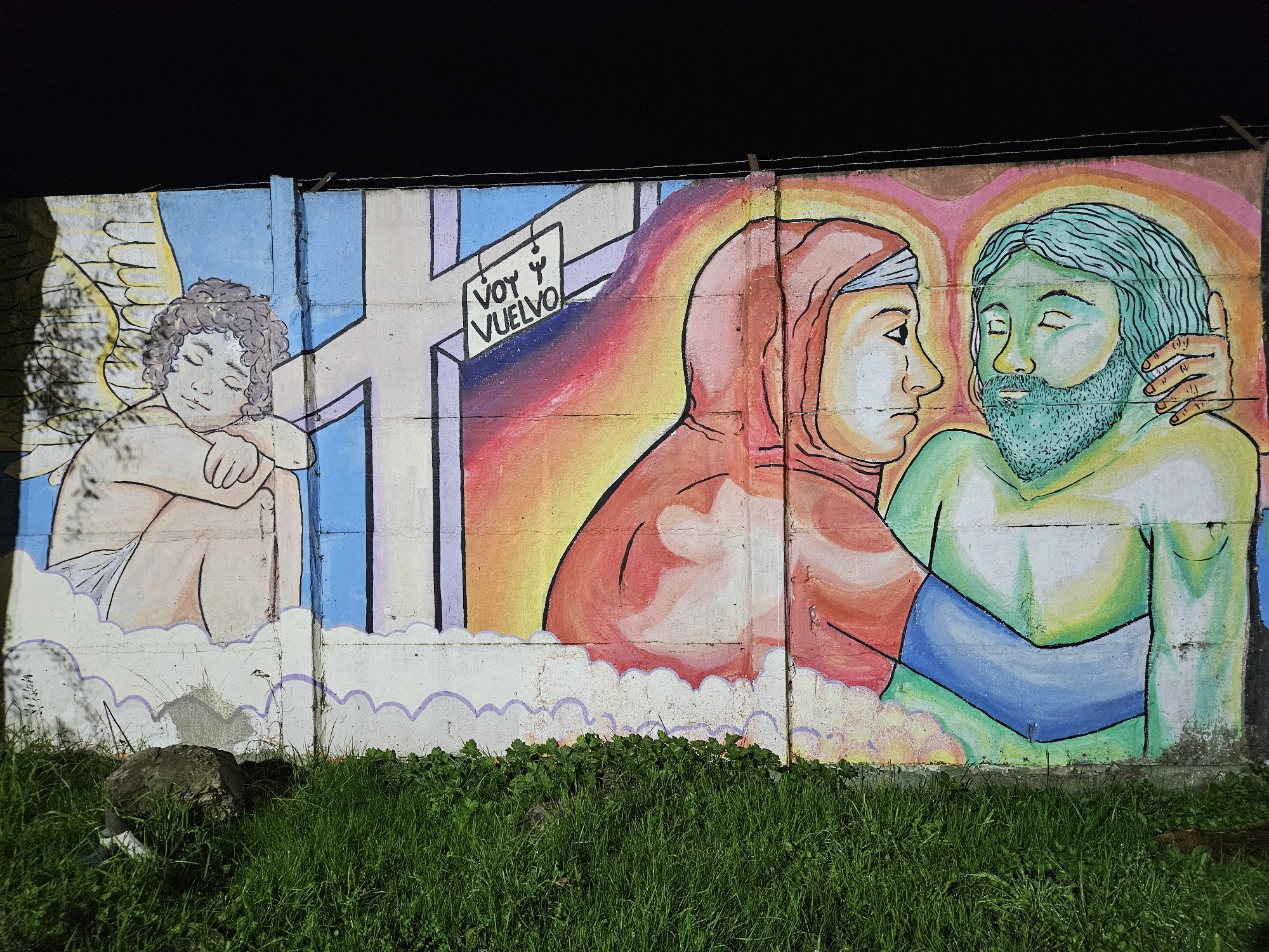 A vibrant street art mural is painted on a concrete wall in Osorno, Chile, depicting a symbolic scene under dark, low-light conditions.
On the left side of the mural, a winged, cherubic figure with curly hair rests peacefully against a light blue background. In the center, a large, stylized cross stands, from which hangs a white sign displaying the Spanish text "VOY Y VUELVO" (I go and I come back). To the right, two figures are in a close embrace against a backdrop of warm, rainbow-like colors. One figure, wearing a red hooded garment, looks towards the other, who has green hair and a beard, closed eyes, and a serene or contemplative expression; their head is cradled by the first figure's hand.
The bottom of the mural features stylized white clouds, and the concrete wall itself shows visible textures and divisions. The ground directly beneath the mural is covered in lush green grass, interspersed with some rocks and debris. The dark sky visible above the wall suggests the image was captured at night or in very dim light. Thin wires run along the top edge of the concrete wall.