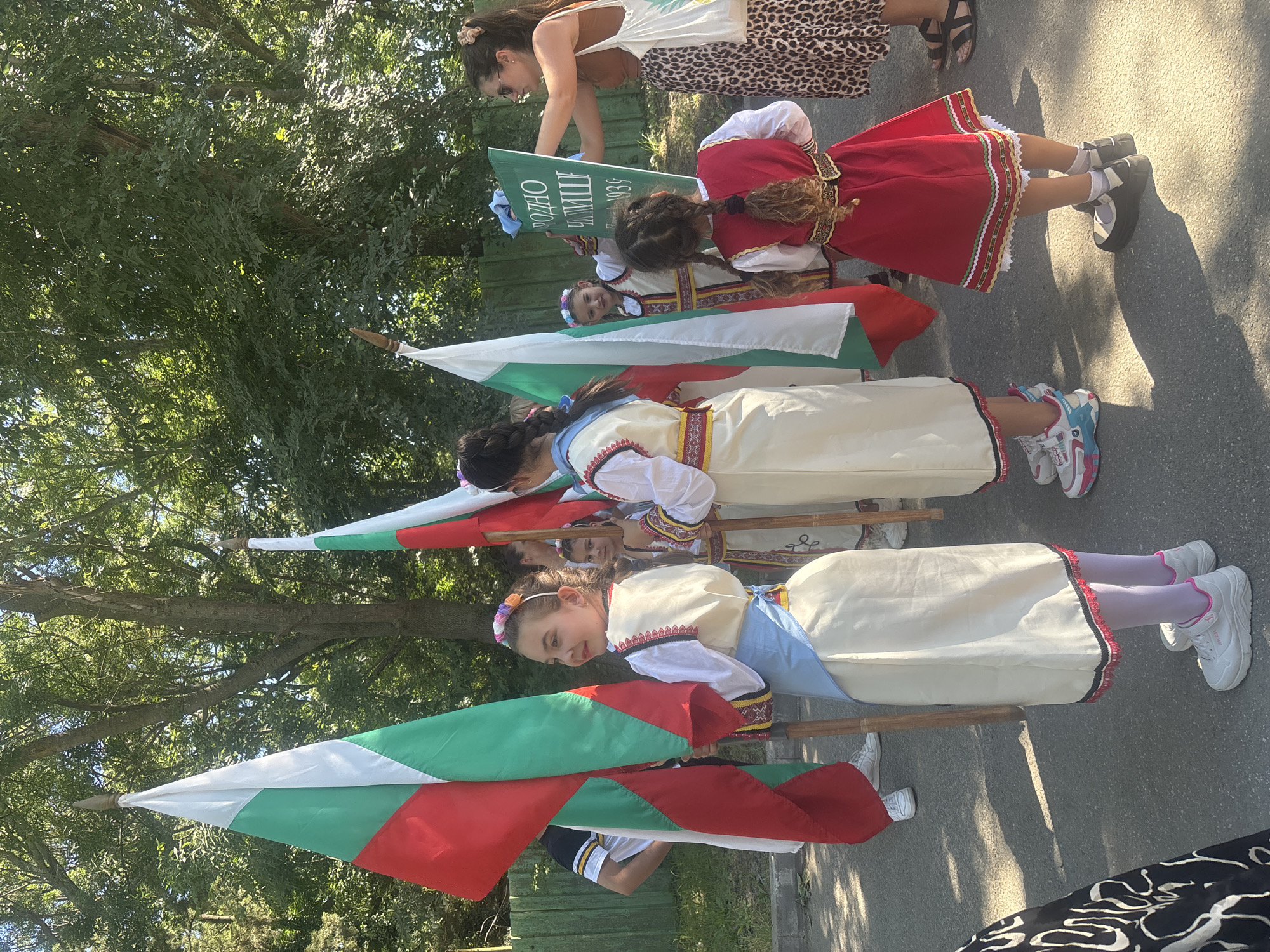 A group of young girls in traditional Bulgarian folk costumes are participating in an outdoor event, possibly a parade or celebration, on a paved street in the city of Burgas, Bulgaria. The girls are holding Bulgarian flags, which are white, green, and red. Some of the girls are wearing embroidered vests and skirts, with colorful headbands. One of the girls is holding a green sign with text written on it in Cyrillic. Behind the children, a woman in a leopard-print skirt and a brown top is holding the sign, and there are trees and a wooden fence in the background. The lighting suggests it is daytime, with shadows cast on the ground. The overall scene conveys a sense of cultural pride and community gathering.