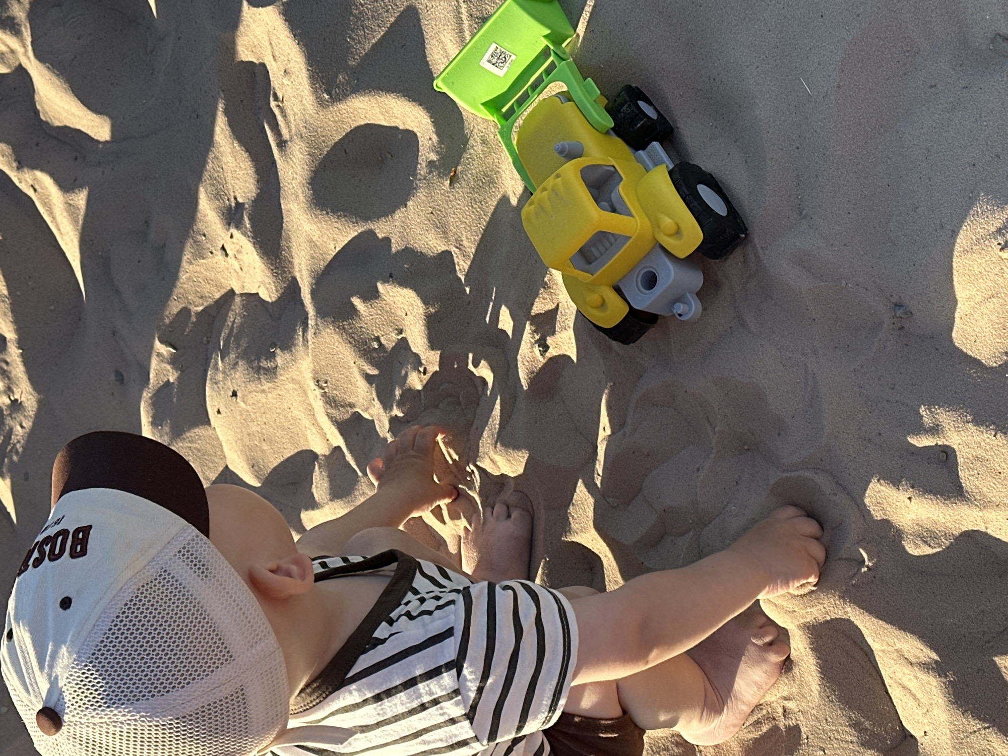 A high-angle shot depicts a young child playing with a toy construction truck in a sand environment. The child, wearing a white and brown baseball cap, a striped shirt, and brown shorts, sits in the sand with legs extended. The cap has "BOST" embroidered on the front. A green, yellow, and gray toy truck sits near the child, casting a shadow on the sand. The sand itself is textured with shadows and light, suggesting a sunny day. The location is a sandy area, possibly a beach, with no additional indications of the city.