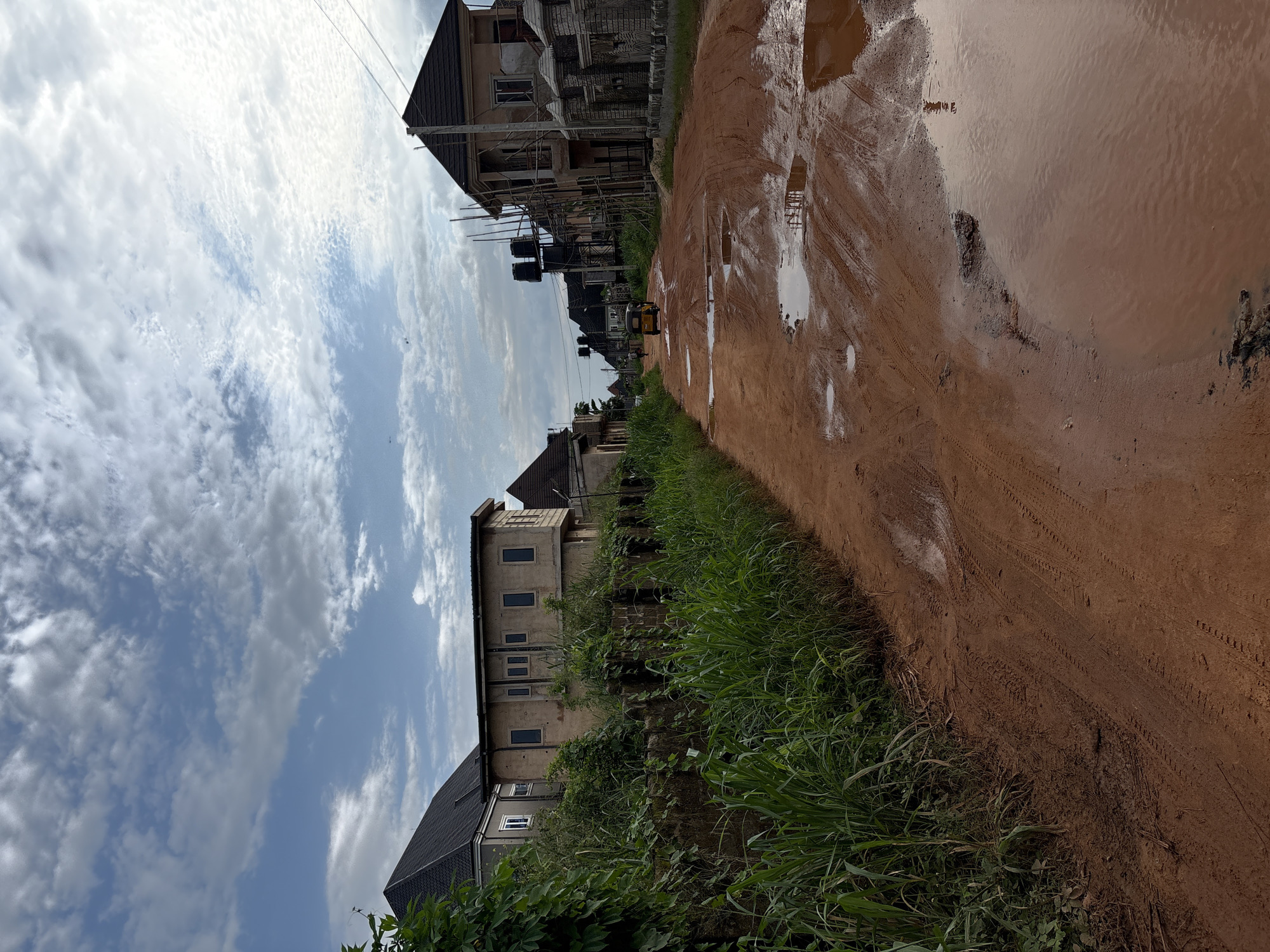 The image depicts a rural road in Obosi, Nigeria, characterized by its reddish-brown, muddy surface with numerous large puddles, indicating recent rainfall. The road is flanked by overgrown green vegetation on the left and residential buildings under construction on the right. Several houses are visible, some appearing partially completed with scaffolding. In the distance, a yellow tricycle, a common mode of transport in Nigeria, is moving away from the viewer. The sky is partly cloudy, with a mix of white and gray clouds against a blue background. The overall scene suggests a developing area with ongoing construction and the challenges associated with unpaved roads in a tropical climate.