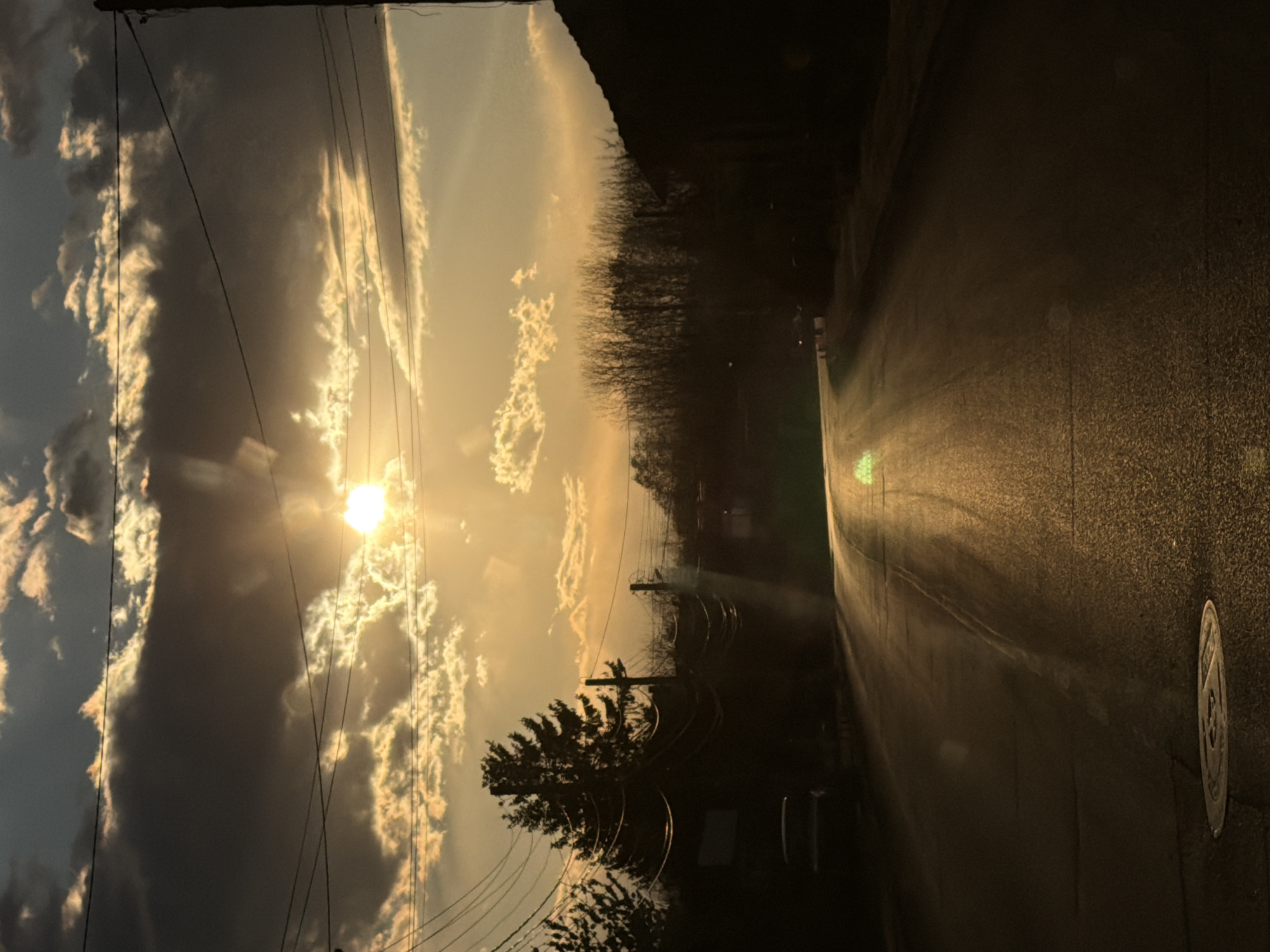 The image depicts a street in Yoakim Gruevo, Bulgaria at sunset. The sun is visible through a break in the clouds, casting a golden light across the sky and the road. Power lines are visible against the sky, and trees line the side of the road. The road itself is dark and wet, reflecting the sunlight. There are no people visible. The scene is still and quiet, conveying a sense of peace.