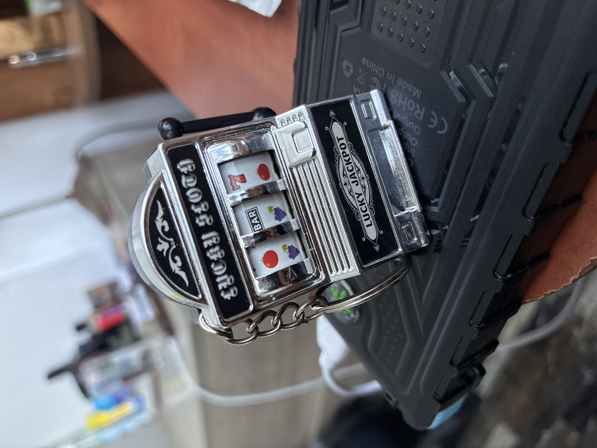The image shows a miniature slot machine keychain attached to the ruggedized case of a smartphone. The slot machine displays the symbols '7', 'BAR', and a tomato, with the words "LUCKY JACKPOT" printed on one side and "LUCKY SLOTH" on the other. The smartphone case has a matte black finish with embossed geometric patterns and labels including "CE ROHS FC Made in China" and input/output information. The background is somewhat blurry, but a table or countertop and some other indistinct objects are visible. There is no information in the image to determine if the image was taken in the city of Varna, Bulgaria.