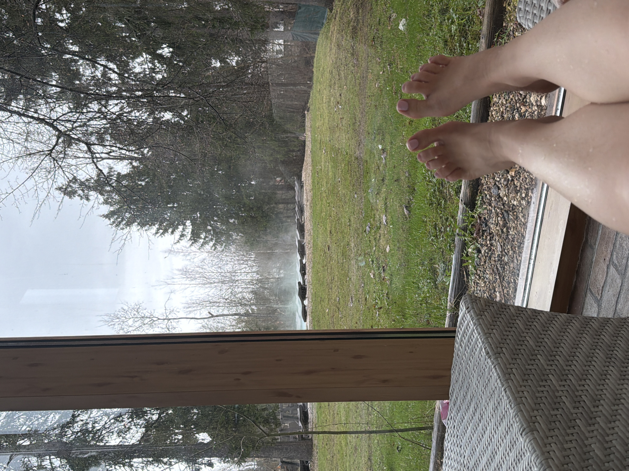The image shows a partial view of a person's legs and feet, resting on a wicker chair or stool. The background consists of a view through a window, depicting a lawn area with green grass, trees, and possibly a body of water or a feature emitting steam or mist. The environment suggests a rural or suburban setting. The location is specified as Dulepovo, Russia, but there are no immediately discernible features in the image that confirm this geographical location. The person is likely relaxing or enjoying the view. The scene gives the impression of relaxation and leisure.