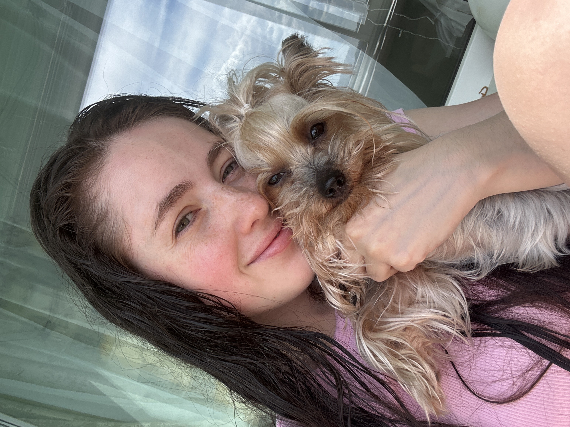 A close-up, overhead selfie shows a young woman with long, dark, wet hair, and light skin with freckles. She is wearing a pink ribbed top. She is holding a small, light brown Yorkshire Terrier dog. The dog is positioned in front of her face, obscuring her mouth. The woman's left arm is around the dog. Her right hand is visible holding the dog's fur. The background appears to be a large window with a reflection of clouds and sky. The lighting suggests it is daytime. There are no discernible landmarks or indicators to suggest a location in Minsk, Belarus.