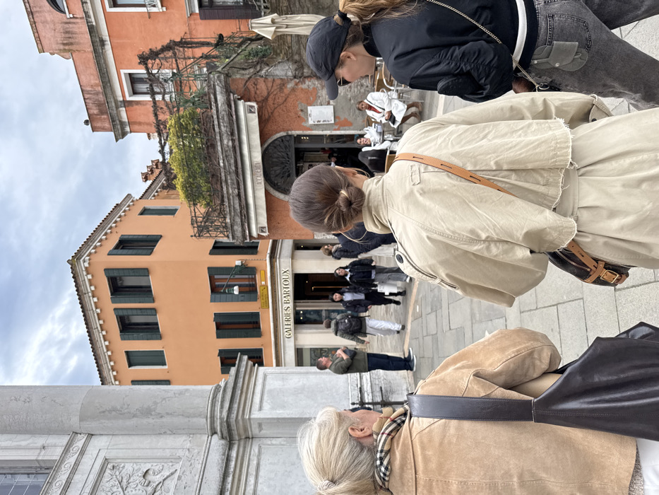 The image captures a bustling outdoor urban scene in Venice, Italy, specifically identified by a street sign as Rio del Malcanton. The sky is overcast, providing diffuse natural light.
In the immediate foreground, three women are observed from behind. The woman on the left has blonde hair in a ponytail, wears a tan suede jacket over a plaid scarf, and carries a dark brown leather shoulder bag. The central woman, with dark hair in a neat bun, wears a beige cropped trench coat and a small brown crossbody bag. The woman on the right wears a black baseball cap, sunglasses, a black bomber jacket, grey jeans, and a black bag with a chain strap and an attached small utility pouch.
The midground features a paved pedestrian area with several individuals. A man in a green shirt stands to the left. A group of four pedestrians walks toward the background. To the right, an outdoor seating area for a café named "ACCADEMIA" is visible, with white tables and chairs. Two individuals are seated there, one in a white coat or robe, and another wearing a red scarf.
The background is dominated by multi-story Venetian architecture. A prominent orange building with dark green window shutters houses "GALERIES BARTOUX" at street level. To its right, another reddish-orange building features an ornate wrought-iron balcony heavily adorned with green foliage and vines, situated above the "ACCADEMIA" establishment. A large, light-colored stone pillar with carved relief details is partially visible on the far left of the frame.