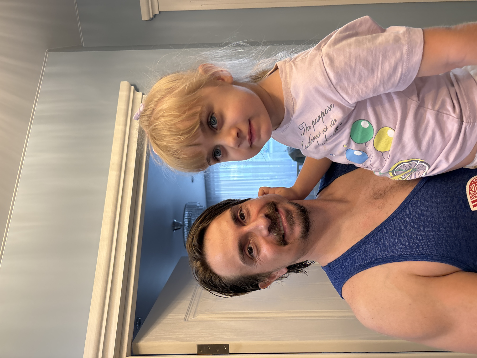 A man and a young girl are posing for a photo in what appears to be a home interior. The man, with brown hair and a mustache and goatee, is wearing a blue tank top and holding the girl. The girl, with blonde hair tied up with a purple bow, is wearing a light pink t-shirt with a graphic of a bicycle and balloons. She is looking towards the camera. The background shows a doorway with white trim, a light-colored wall, and a glimpse of a chandelier in a dimly lit room beyond the doorway. There is no indication of the city of Sinyavino, Russia, in the image.