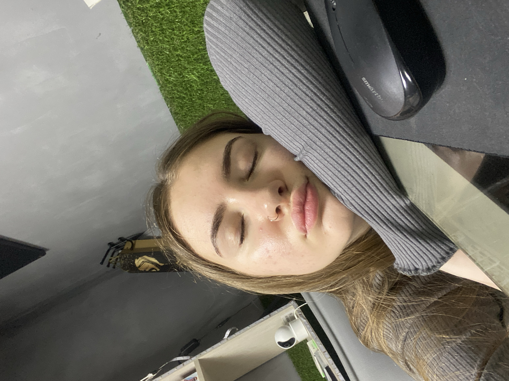 A young woman is captured in a close-up, selfie-style shot, with her eyes closed and head resting on her arm, suggesting she is sleeping or napping. Her face is turned slightly towards the camera. She is wearing a grey, ribbed garment on her arm, which is supporting her head. In the foreground, a black computer mouse is visible on a dark mousepad. The background reveals a grey wall with a textured finish, a section of artificial green turf, and a shelf with a small white camera device. The setting appears to be indoors, possibly a home office or study area. The image is taken from a slightly elevated angle. There are no other individuals visible in the frame. The location is identified as Dyakovo, Russia.