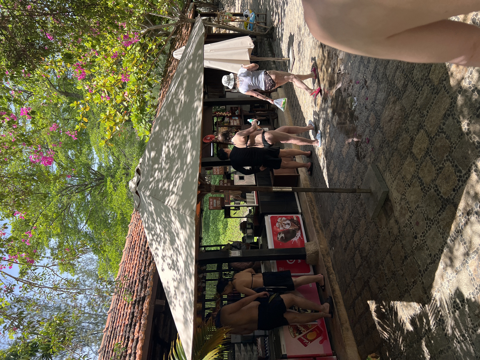 The image depicts an outdoor scene at a seaside establishment in Tay Nha Trang Ward, Vietnam. In the foreground, a portion of a person's bare shoulder and upper arm is visible, suggesting the photographer is part of the scene. The middle ground features a shaded area under a large umbrella, where a small open-air shop or kiosk is set up. A long, tiled counter is visible, with several ice cream freezers and display cases. Behind the counter, a person can be seen serving customers. In front of the counter, a man and a woman, both wearing swimwear, are interacting. The man is shirtless, wearing black shorts, and the woman is wearing a black bikini. Further to the right, another woman in a white t-shirt and patterned bikini bottoms walks away from the kiosk, carrying a plastic bag. She is wearing a hat and pink flip-flops. The ground is paved with cobblestones. Lush green trees and some flowering pink bougainvillea are visible in the background, along with a tiled roof of a building. The overall impression is a casual, sunny day at a beachside resort or attraction.