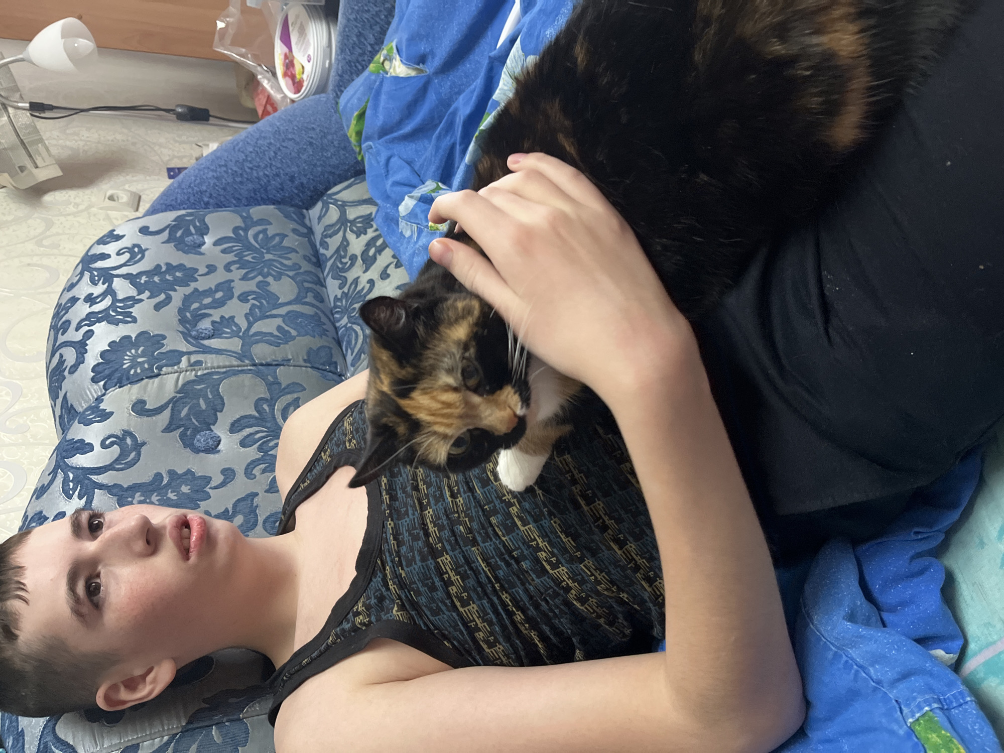 A young person lies on a blue floral-patterned couch. A tortoiseshell cat is on the person's chest. The person is petting the cat. The person is wearing a dark tank top. A blue blanket is nearby. A lamp and other objects are visible in the background. The location is Chaikovsky, Russia.