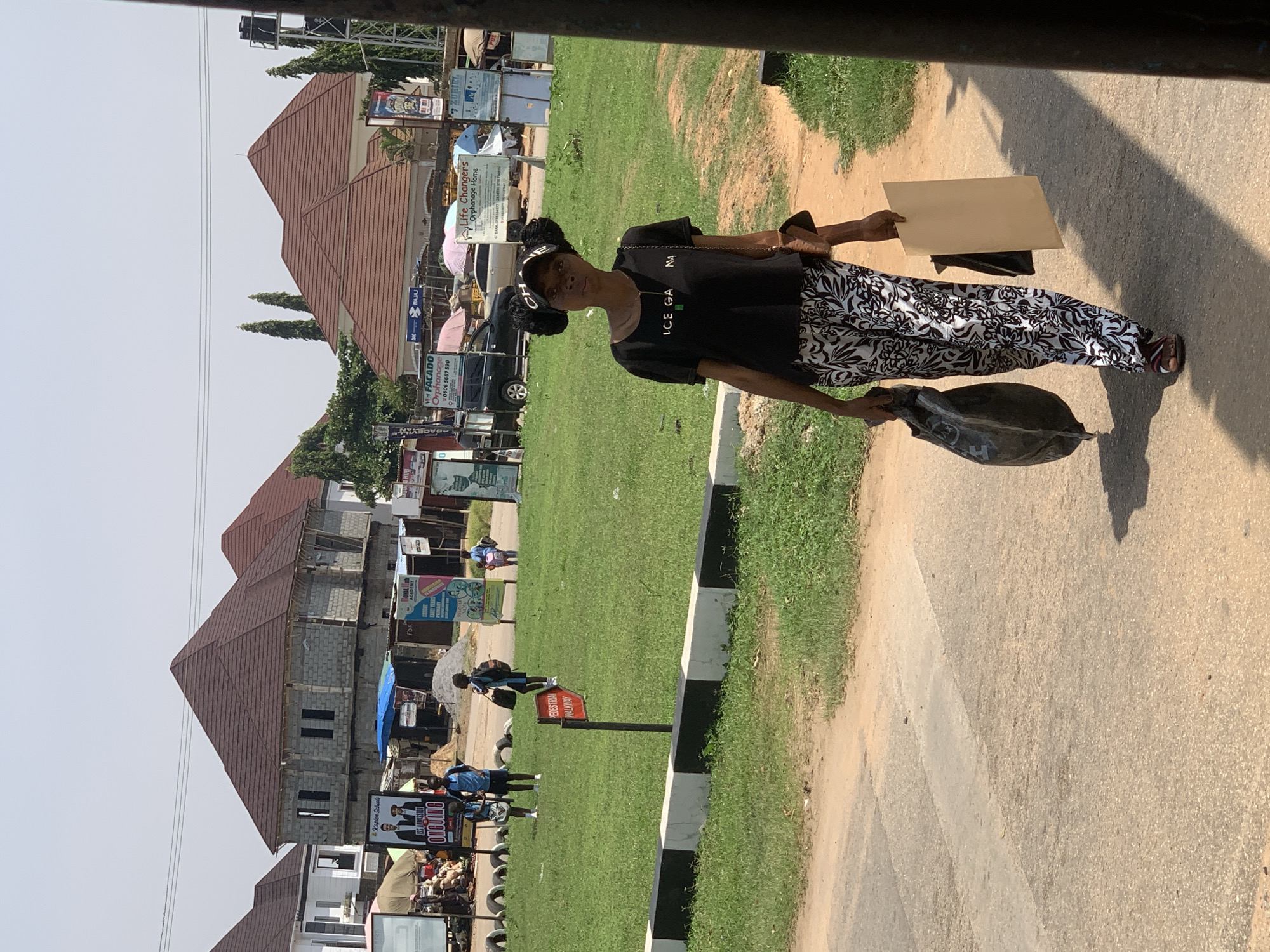 A young Black woman walks along a paved road on a bright, sunny day in Abuja, Nigeria. She is wearing a black t-shirt with the words "ICE GA NA" in white lettering, a black baseball cap, and patterned black and white pants. Her hair is styled in two large buns on top of her head. She carries a large black trash bag in her left hand and a flat, light brown paper bag in her right hand. Her feet are clad in sandals. The woman is walking away from the camera, towards a grassy area with several buildings and signs in the background. The buildings appear to be under construction or recently completed, with reddish-brown roofs. Among the signs are advertisements for schools and an orphanage. Several other people, including children, are visible in the distance on the grassy area. The overall environment suggests a developing urban or suburban setting.