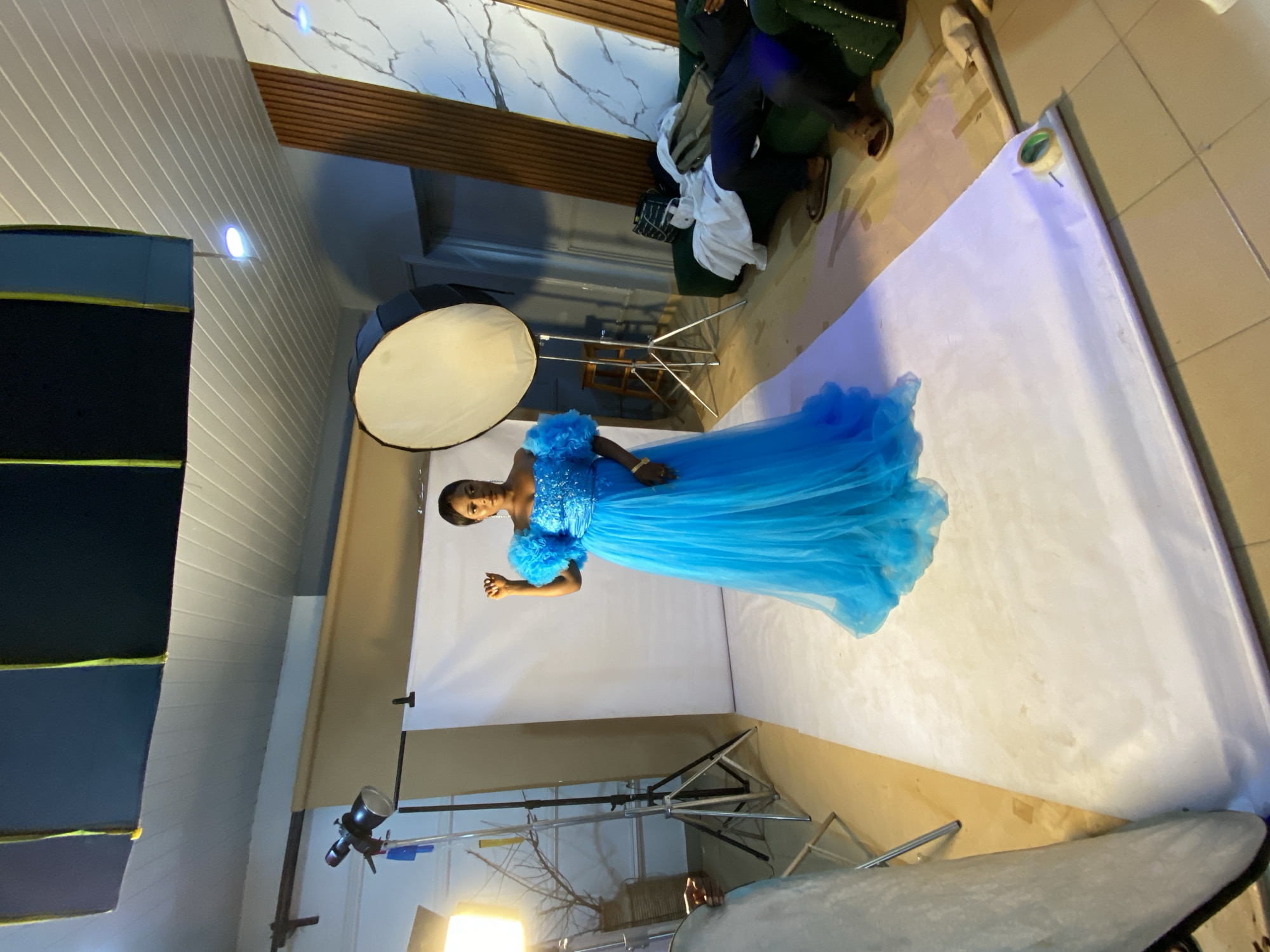 This media file captures a studio photoshoot scene in Bukuru, Nigeria. The primary subject is a young Black woman dressed in a voluminous, bright blue gown with intricate embellishments on the bodice and ruffled sleeves. She is posing on a white backdrop that extends to the floor, with a large softbox light positioned to her left, creating a soft, diffused illumination.
In the background, a few people are visible. To the upper right, two individuals are seated on a dark green couch. One person appears to be wearing dark trousers and sandals, and their legs are crossed. Another individual is wearing dark trousers and a light-colored shirt, with a white garment draped over their lap. A black bag is placed on the floor near them.
The setting appears to be an indoor studio. The walls are decorated with a white, marbled wallpaper interspersed with wooden slats. To the left of the frame, a portion of a studio backdrop structure with dark blue and black panels is visible, along with a ceiling light fixture. In the foreground, studio lighting equipment, including a light stand with a visible bulb, is present. The floor is tiled with light brown tiles.
The overall atmosphere suggests a professional or semi-professional photoshoot. The lighting indicates it is likely daytime or a well-lit indoor space, with no indication of weather conditions. The subject's pose and attire convey a sense of elegance and poise.