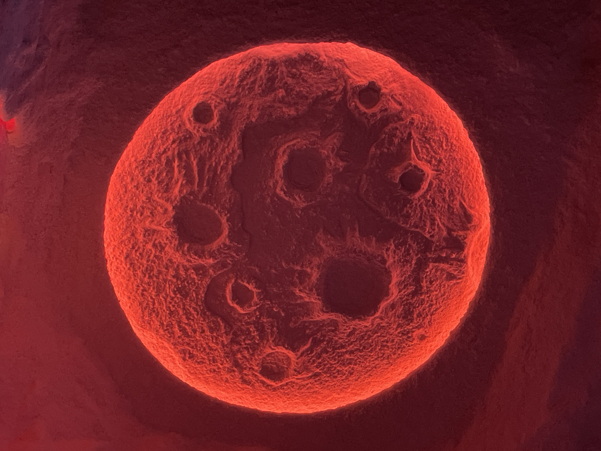 The image depicts a highly magnified, spherical, reddish-orange object with a rough, cratered surface. The object is centered within a red-toned environment. The uneven surface suggests textures and potential indentations. There are no discernible people, actions, or interactions within the frame. The location cannot be determined from the visual data.