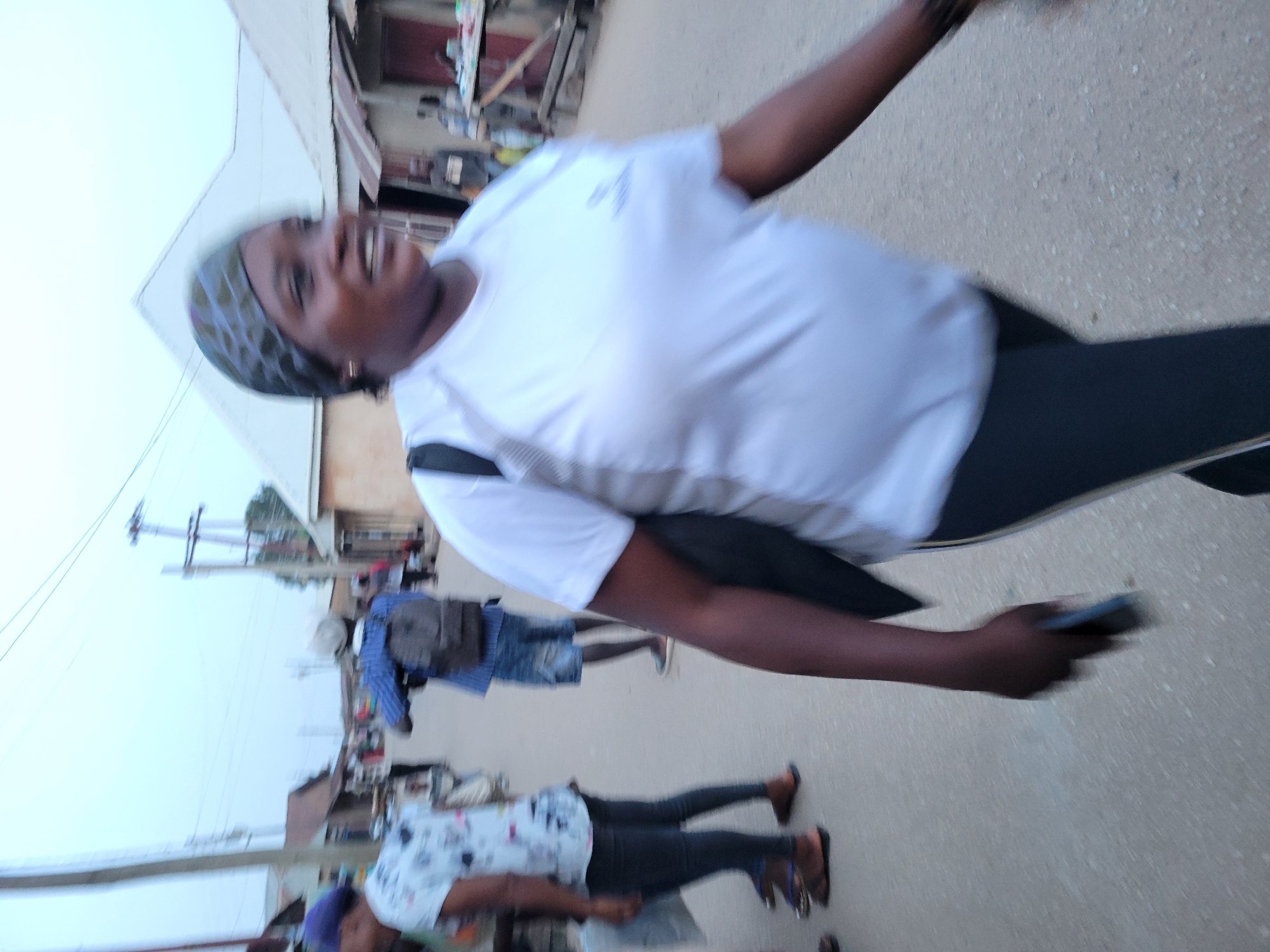 The image, taken in what appears to be a residential area in Jos, Nigeria, is blurred due to motion. A woman is in the foreground. She is wearing a white t-shirt, dark leggings, and a patterned headscarf. She has a dark complexion and is smiling. She is holding a cell phone in her right hand.
Behind her are other individuals walking on the street. One person is wearing a blue shirt and denim shorts with a messenger bag. Another is wearing a patterned shirt and black pants.
The environment includes buildings with corrugated metal roofs and power lines. The ground is paved. The lighting suggests it is daytime.