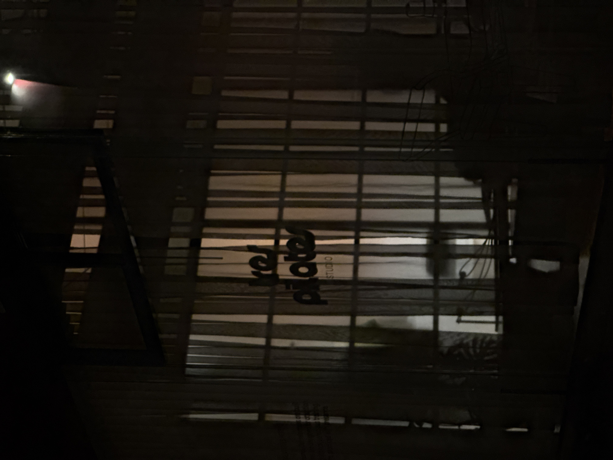 The image shows an aerial view of a street in Puebla, Mexico at night. The street is characterized by horizontal lines, which appear to be shadows cast from an overhead structure, possibly a bridge or a large sign. There is a sign for "Arter Studio" visible. The environment is dark, with a minimal amount of lighting from distant sources. There are no discernible people visible in the image.
