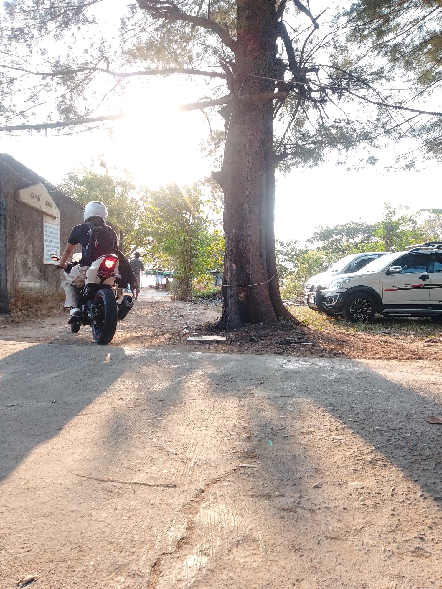 In Cikal, Indonesia, the image captures a scene with a person on a motorcycle, viewed from behind, moving away from the camera on a dirt road. The person is wearing a white helmet and a dark t-shirt, with a backpack on. The motorcycle is white and black with a red taillight illuminated. To the left of the road, a building with a sign is visible, and further back, a second person walks away from the camera. To the right of the road, two white SUVs are parked. The environment is characterized by a large tree trunk in the foreground, with its branches extending into the bright, sunlit sky. The ground is a mixture of dirt and some paved sections, with shadows cast by the tree. Lush green foliage is present in the background, suggesting a semi-rural or coastal setting.
