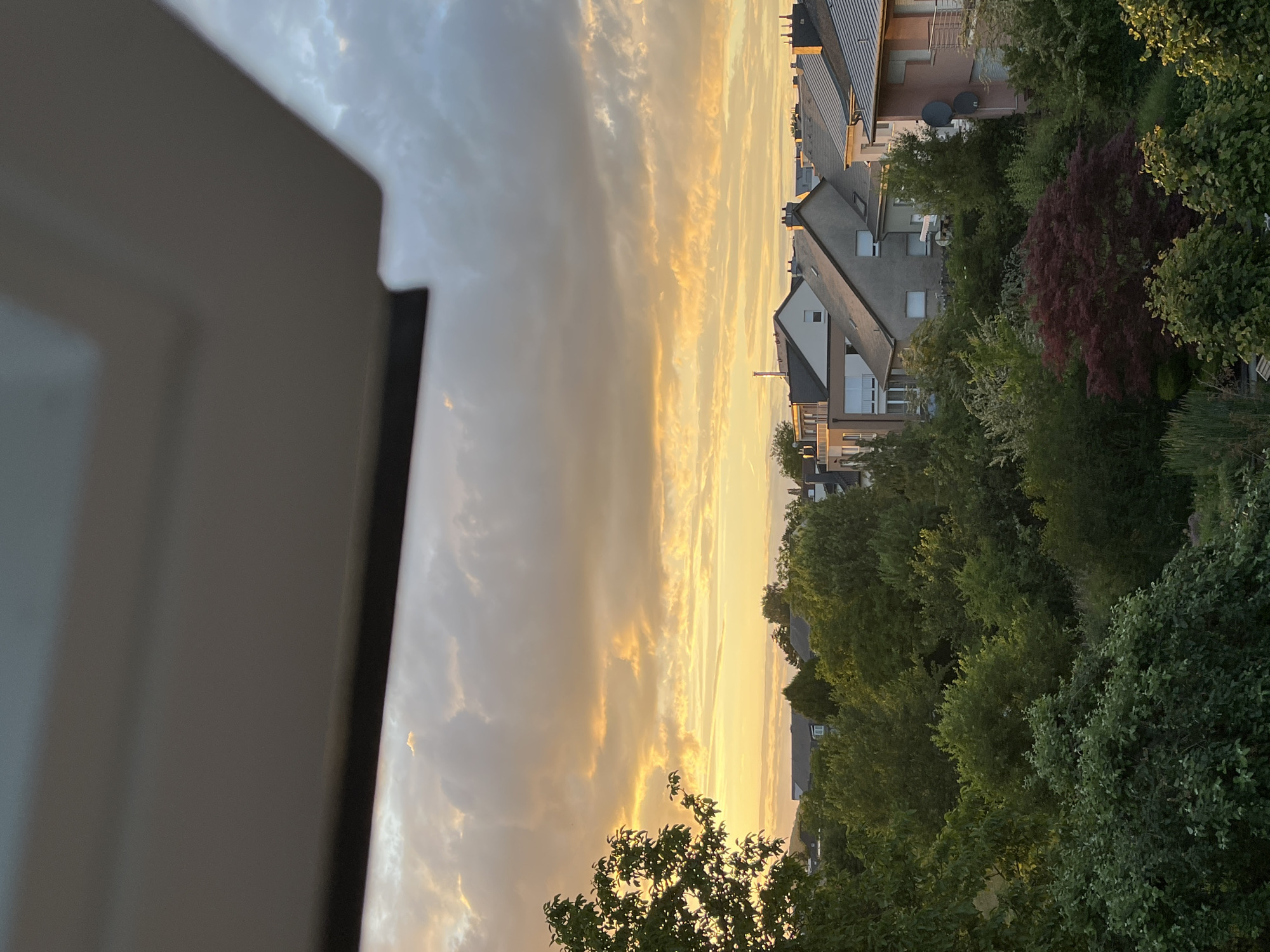 The image depicts a suburban landscape at dusk in Mondercange, Luxembourg. A substantial overhang, likely part of a building's structure, occupies the foreground, casting a shadow over the upper portion of the scene. Below this, a panorama of residential rooftops and lush greenery unfolds. The sky is a canvas of dramatic clouds, illuminated by the warm, golden hues of the setting sun, with patches of blue visible. The architecture appears to be modern, with pitched roofs and various building materials. Dense trees and gardens fill the spaces between the houses, suggesting a well-developed residential area with significant green spaces. No individuals or overt actions are captured within the frame.