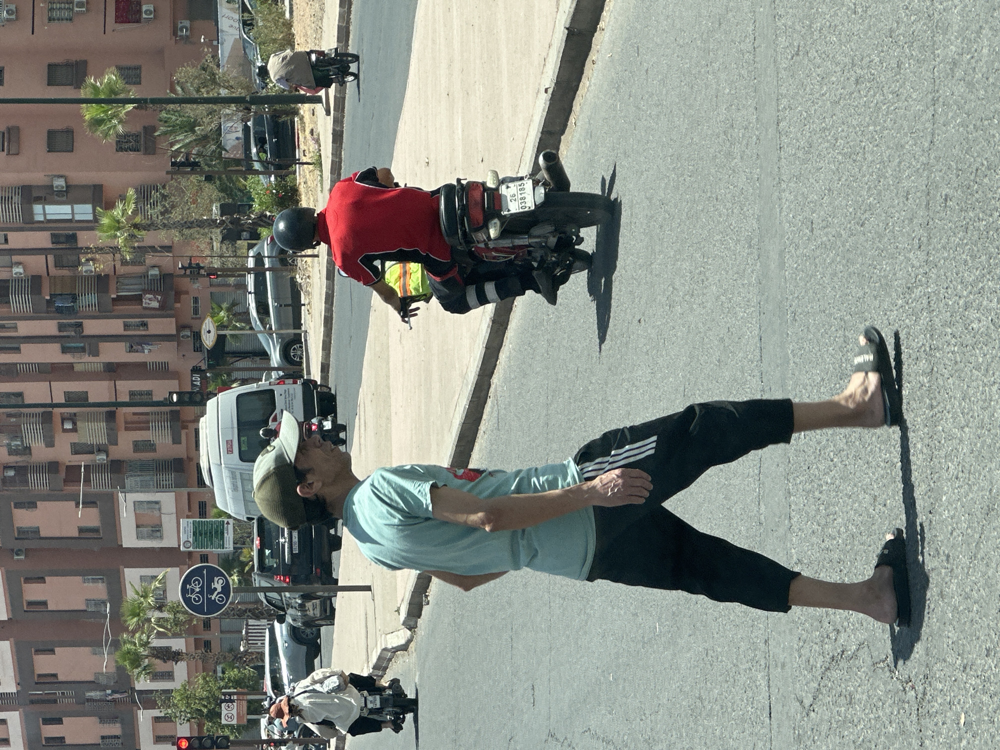 The image depicts a street scene in Marrakesh, Morocco, with a man wearing a light blue t-shirt, dark pants with white stripes, and a green cap walking on the right side of the road, wearing black sandals. A motorcycle rider in a red shirt, black helmet, and dark pants is driving on the road. The motorcycle has a license plate visible as "26-038765". Traffic is present, including a white van, dark-colored cars, and another motorcycle with a rider in white, visible in the background. A bike/motorcycle lane sign is posted on the side of the road. The background features multi-story buildings with multiple windows and air conditioning units, as well as palm trees.