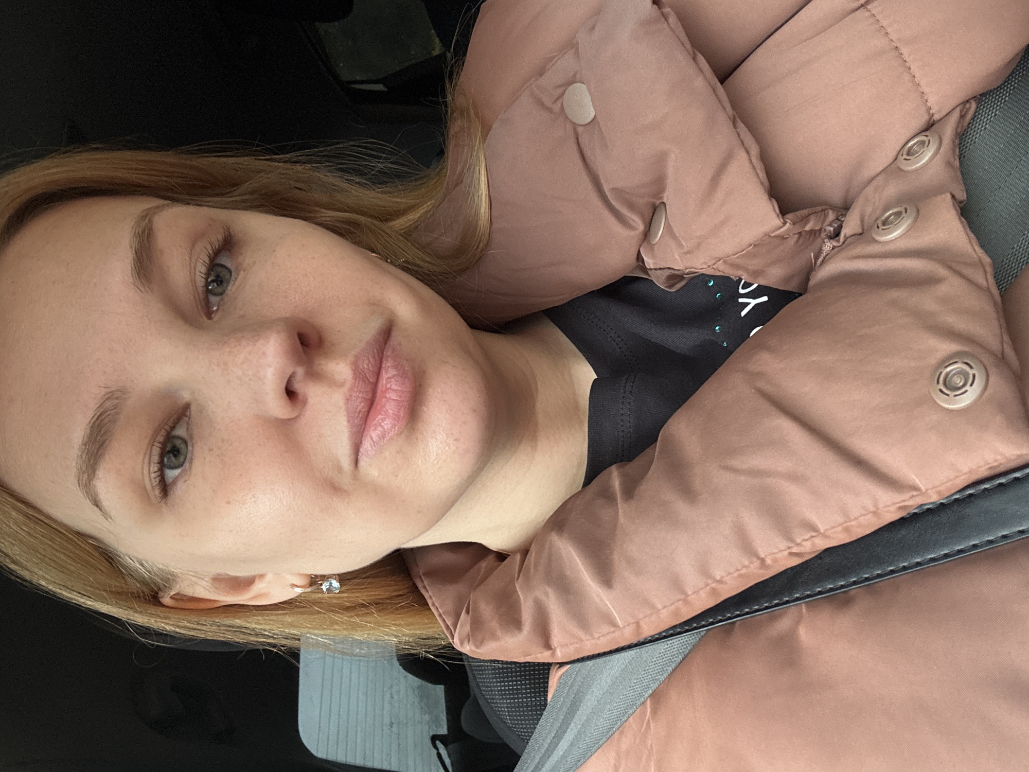 A light-skinned woman with blonde hair is seated in what appears to be a car. She is wearing a brownish-pink puffer jacket over a black top. A seatbelt is visible. She has freckles on her face and is wearing an earring. The background is dark, suggesting it could be inside the vehicle. There is no information given in the image to suggest that it is in Rusanovo, Russia, or any other geographical location.