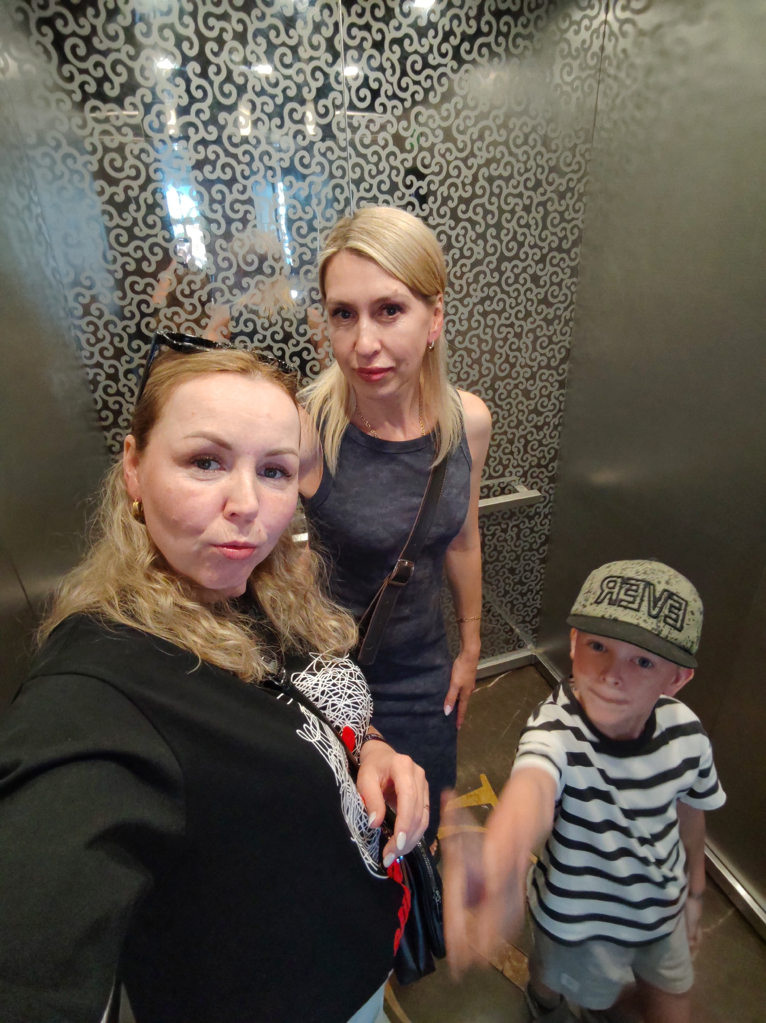 The image depicts three individuals inside an elevator. The environment is characterized by mirrored walls with decorative patterns and metal fixtures.
A woman with blonde, curly hair, wearing a black t-shirt with a white design, is positioned in the foreground, taking the photo. Behind her stands another woman with straight, blonde hair, wearing a dark gray dress and a brown shoulder bag. To the right is a young boy wearing a striped shirt, gray shorts, and a green cap with a word backward on it. He is pointing towards the camera.
The scene is identified to be in Alanya, Turkey, based on the reflection in the mirrors revealing the city's cityscape.