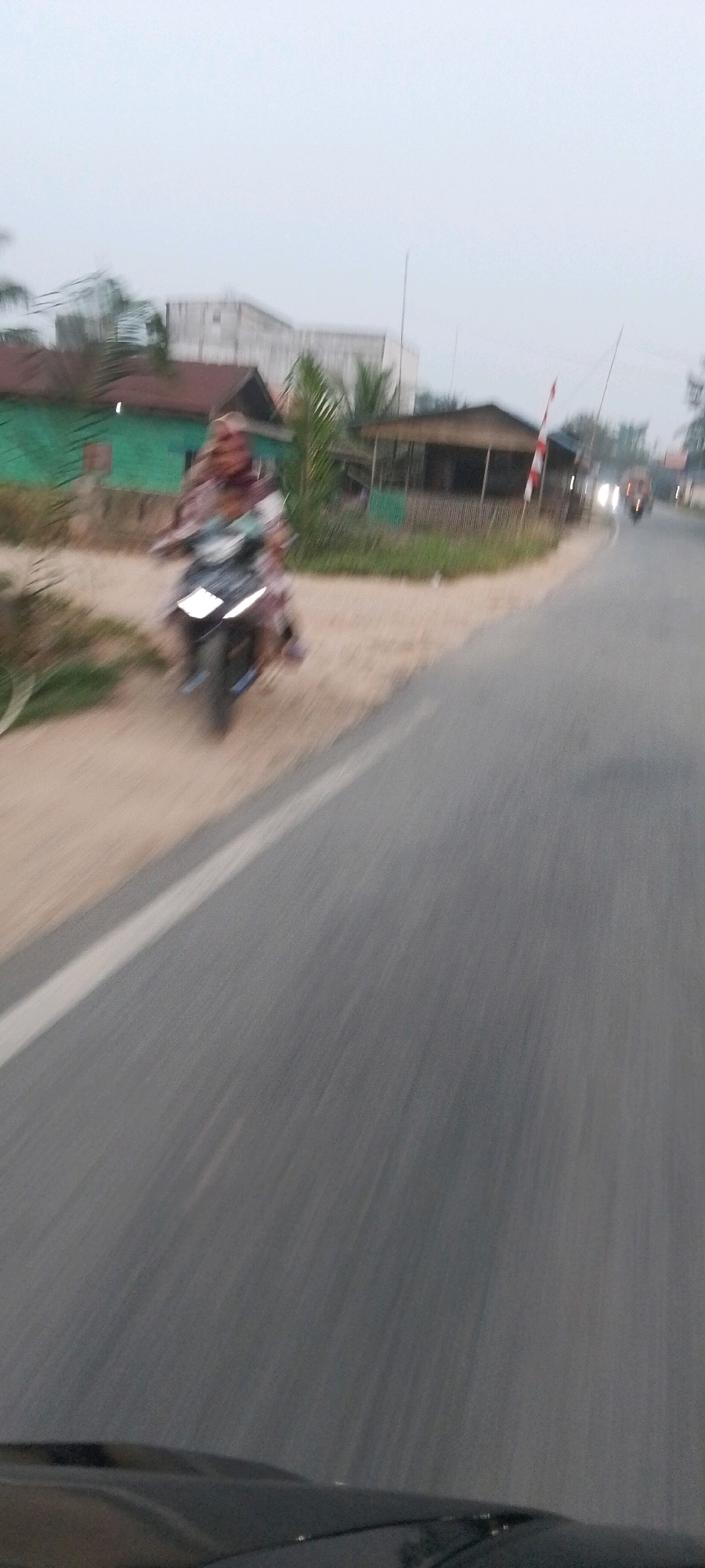 The image captures a motion-blurred scene on a dusty road next to a paved road in Pematang Sikek, Indonesia. In the foreground, the top of a dark-colored vehicle is visible. On the left side of the frame, a person is riding a motorcycle with another person behind them. The motorcycle is dark-colored with bright headlights. The riders are wearing light-colored clothing. Behind the motorcycle, there are some low-lying buildings with green and brown roofs, as well as palm trees and other vegetation. To the right of the paved road, there is a small hut-like structure with a brown roof and bamboo walls, and a red and white striped pole, possibly part of a barrier. Further down the paved road, two vehicles with illuminated headlights are approaching. The sky is overcast with a light gray hue. The overall impression is of a rural or semi-rural area during daylight.