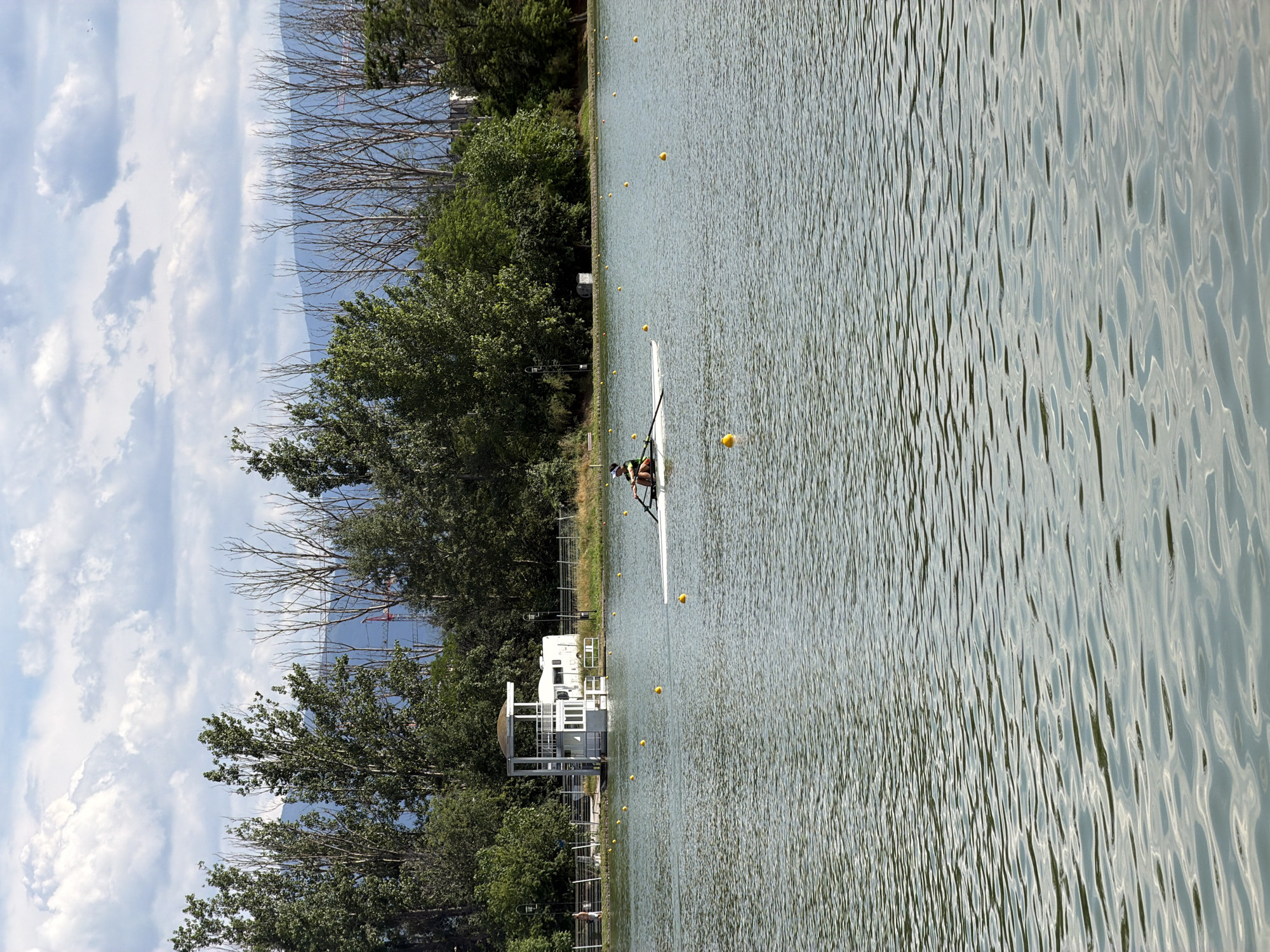 The image shows a rowing scull on a body of water, likely a lake or river. A single person is in the scull, rowing. The surrounding environment includes trees along the bank, a white building with a tower, and a large white vehicle, possibly an RV, parked nearby. In the distance, there are hills or mountains and a cloudy sky. Yellow buoys are scattered on the water, marking a course. The scene suggests a sporting event or practice session taking place outdoors. This location is likely the Plovdiv Rowing Canal, a venue known for hosting rowing competitions and training in Plovdiv, Bulgaria.