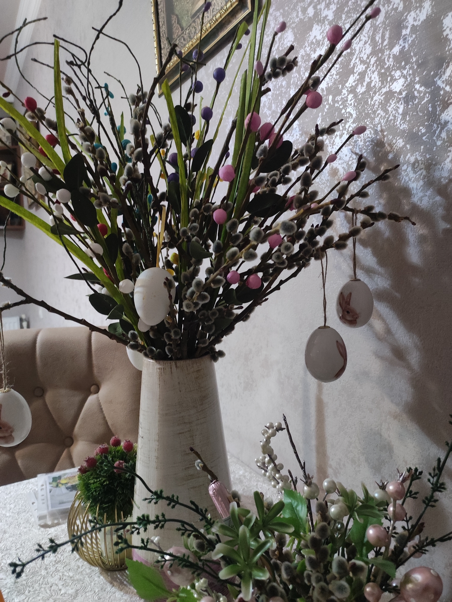 An indoor still life composition features a tall, cream-colored vase filled with branches adorned with fuzzy catkins, green leaves, and small, colorful decorative spheres in shades of pink, blue, and purple. Several white Easter eggs, some decorated with faint pink bunny figures, are suspended from the branches by thin twine. To the left of the vase, a tufted, light brown upholstered chair is partially visible. On the table, to the left of the vase, a small green plant in a gold wire basket sits on a white, patterned tablecloth. To the right of the vase, another arrangement of faux greenery, pink berries, and white beads is visible. The background consists of a textured, light-colored wall. The scene appears to be a domestic setting, likely decorated for Easter, and is not specific to Akhalkalaki, Georgia.