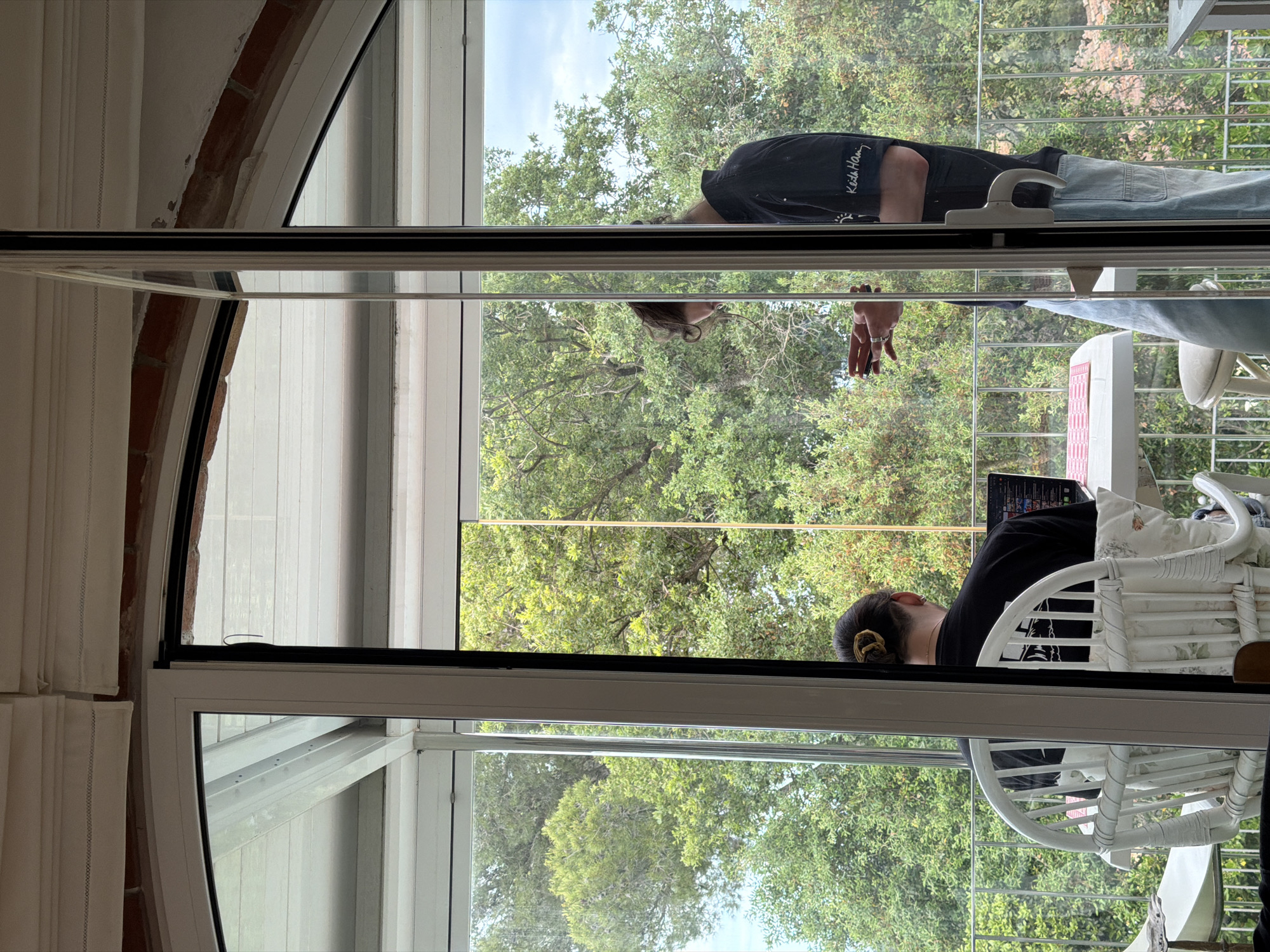 The image shows a person looking through a window in Pals, Spain. The person is wearing a black shirt and light blue jeans and appears to be on a balcony. Inside, another person is working on a laptop while seated on a white wicker chair. A white desk is positioned in front of the laptop and chair. The window frame is white with a black trim. The window appears to reflect the outside environment, including green trees. The building appears to be constructed of brick and white walls.