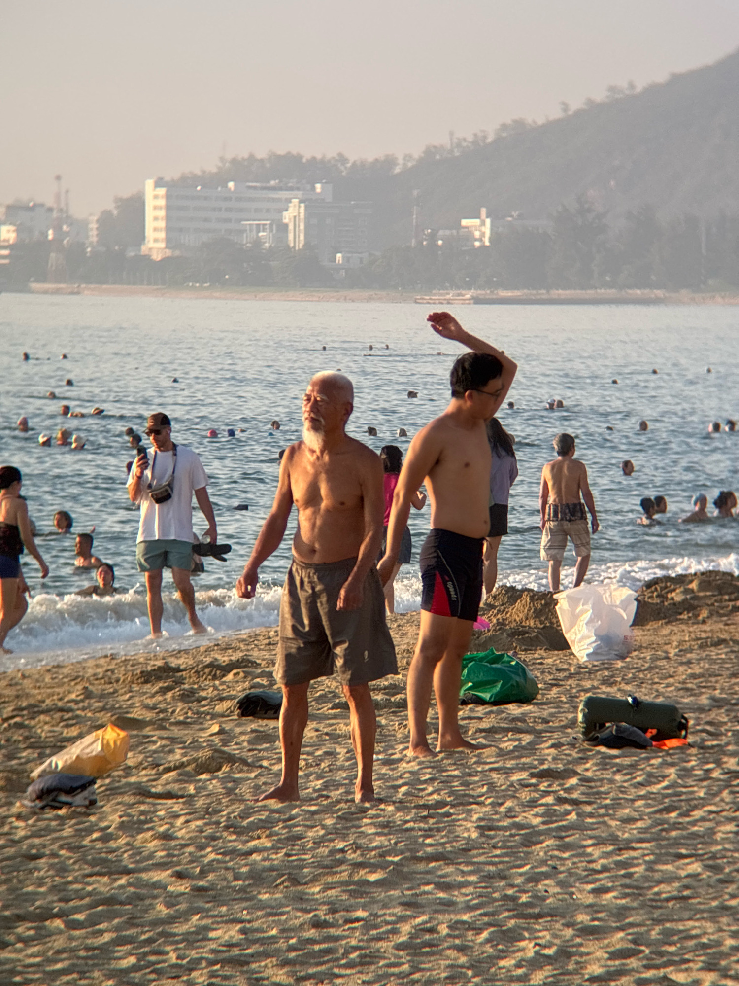 The image depicts a beach scene in Nha Trang, Vietnam, during what appears to be late afternoon or early evening, with a hazy sun casting a warm glow. The foreground is dominated by sandy beach with several objects including a green bag, a white plastic bag, and a rolled-up dark green object with an orange strap. In the mid-ground, two elderly men are standing near the water's edge. The man on the left is shirtless, wearing grey shorts, and has a white beard. The man on the right is also shirtless, wearing black and red swim trunks, and has his left arm raised. Behind them, numerous people are visible in the shallow water and on the beach. Some are swimming, while others are standing or walking. In the background, the ocean stretches out towards a coastline with buildings and a hillside covered in vegetation. The water is calm with gentle waves reaching the shore. The overall atmosphere suggests a relaxed, communal beach setting.