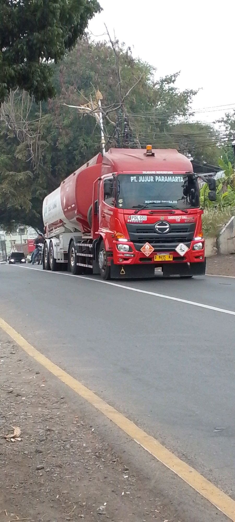 A red Hino tanker truck with a white and red cylindrical tank is driving on a paved road, with a yellow line dividing the lane. The truck has "PT. JUJUR PARAMARTA" printed in white on the windshield. There are hazard symbols on the front of the truck. In the background, there are trees and some power lines. A black car is visible in the distance on the left side of the road, and a person on a motorcycle is also present nearby. The scene appears to be a street in Cilaku, Indonesia.