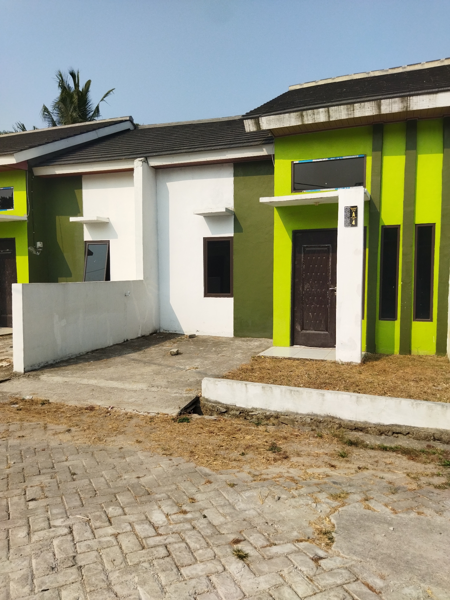 This is a medium-angle, eye-level exterior shot of a row of modern houses under a clear blue sky, located in the Suka Sari area of Indonesia. The houses feature a combination of white and bright lime green exteriors. The house in the foreground on the right has a lime green facade with vertical darker green stripes, a dark brown front door, and three tall, narrow, dark-tinted windows. A white pillar supports a small overhang above the door. A sign with the letter "A" and the number "4" is affixed to the white pillar. The house to the left of it is primarily white, with sections of lime green. It has a dark-tinted window on the left side and another on the right side of its white section. A low white wall extends from the front of this house. In front of the houses, there is a paved area made of interlocking rectangular bricks, with patches of dry grass and dirt. The overall scene depicts a newly developed residential area, possibly a housing complex.