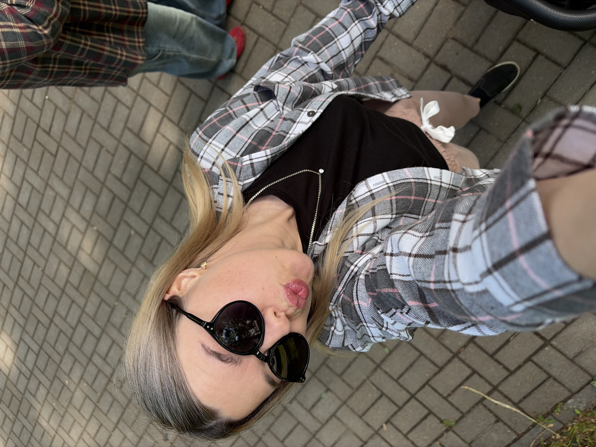A blonde woman is the central subject of this high-angle selfie, captured outdoors on a paved surface. She is seen from the chest up, with her head tilted, looking down at the camera, and her lips pursed in a "kiss" face. She wears dark, round sunglasses and a small gold-colored stud earring. Her attire consists of an open plaid shirt featuring grey, white, black, and pink checks, worn over a black t-shirt. A silver necklace with a small clear gem adorns her neck. Light brown or beige pants/shorts with a white drawstring are visible below her t-shirt.
The background shows the legs of another individual wearing blue jeans and a dark plaid shirt (dark blue, red, and black checks) with red shoes. A black shoe, possibly belonging to the woman or a third person, is also visible. The ground is a paved pathway or sidewalk, composed of grey rectangular pavers laid in a herringbone pattern. Small patches of grass and weeds are present along the edges of the path. A black, textured object resembling a stroller handle is partially visible in the upper right corner.
The activity taking place is a casual selfie, with the woman posing playfully. The scene is set outdoors during the daytime, indicated by natural lighting, though the exact weather conditions are not fully clear, the sunglasses suggest brightness. There is no visible text in the image. The location context is Chaikovsky, Russia.