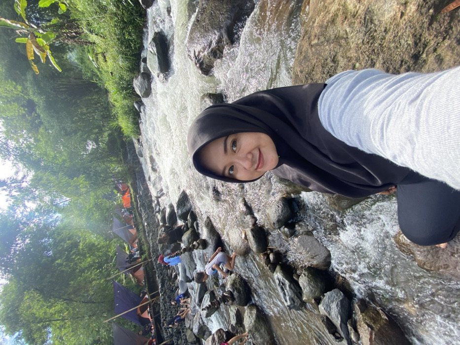 A woman in a black hijab smiles at the camera, positioned near a rocky stream. Flowing water rushes around her, reflecting light. A camping area with tents is visible in the background, surrounded by lush green trees. People are gathered near the stream, some appearing to play in the water. The camera angle suggests the photo may be a selfie.