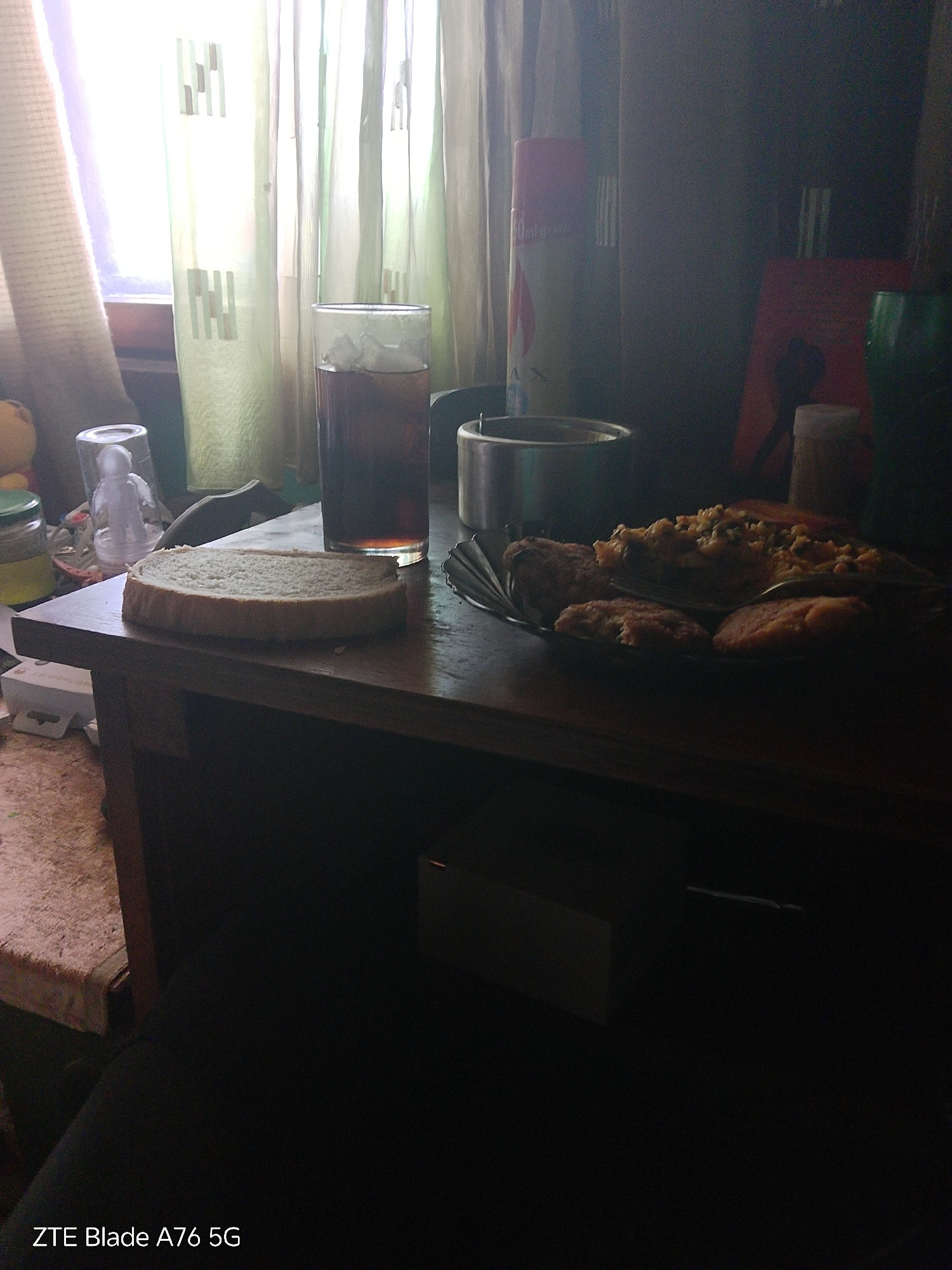 The image, captured by a ZTE Blade A76 5G device, depicts an indoor scene in the city of Stara Zagora, Bulgaria. The primary light source originates from a window in the background, resulting in strong backlighting that silhouettes foreground objects and overexposes the curtained window area.
On a dark wooden table occupying the mid-ground, a thick, oval slice of white bread rests centrally. To its immediate right, a clear glass contains a dark liquid and multiple ice cubes. Further right, a dark plate or bowl holds a serving of a mixed dish, appearing to be scrambled eggs or a similar preparation with visible particulates, accompanied by three rounded cookies or small cakes. A spoon or fork is partially visible on the plate.
Behind the food items, a silver-colored metallic bowl or pot is present. To its left, a red and white aerosol can, partially labeled "400ml grati...", is discernible. Further to the right and largely obscured by shadow, a red rectangular object, a small white cylindrical shaker-type container, and a dark green bottle are situated.
To the left of the bread slice, partially visible items include a glass jar with a yellow lid and contents, and a small clear container housing a white, stylized figurine, possibly an astronaut.
The background reveals a window covered by two layers of curtains. The inner layer is a light green, semi-sheer fabric featuring a pattern of vertical rectangular bars. The outer curtain, positioned on the left, appears to be a beige or off-white opaque material. Sunlight intensely illuminates these curtains. The lower left corner of the image shows a segment of a textured floor or surface beneath the table, possibly a light-colored, patterned textile. No individuals are present in the scene, and no specific actions or interactions are depicted beyond the arrangement of objects on the table.