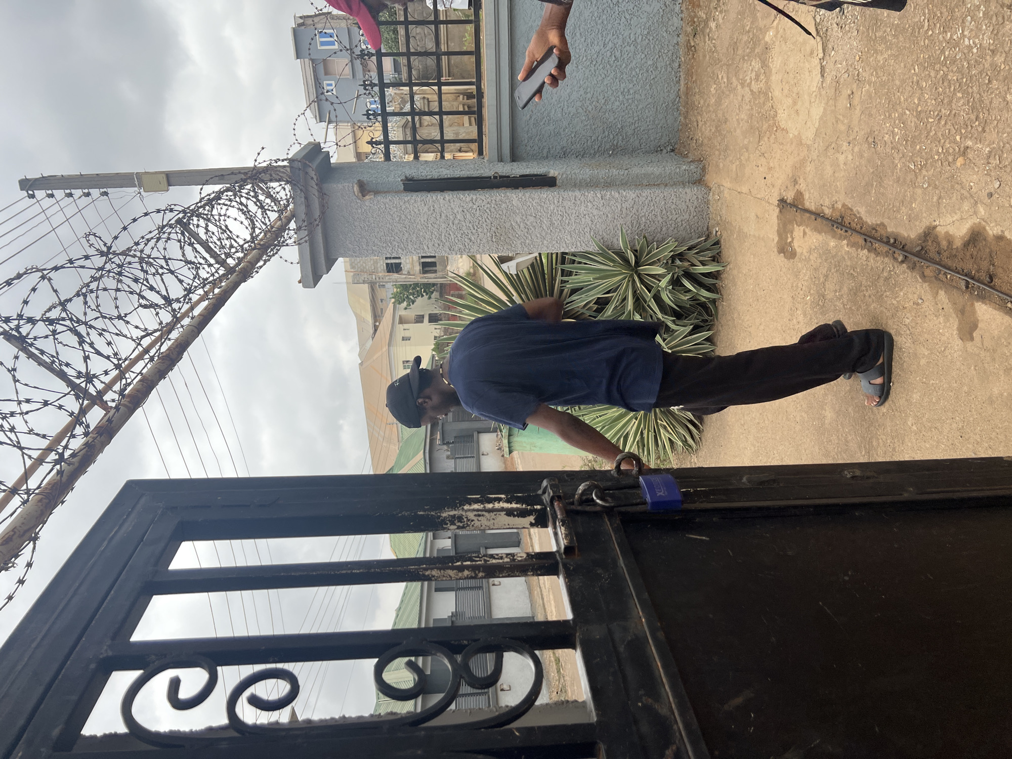 This image, taken in Abuja, Nigeria, depicts an outdoor scene at the entrance of a property on an overcast day.
In the center, a person stands facing away from the camera, wearing a dark blue t-shirt, dark trousers, a black baseball cap, and grey sandals. Their left hand is near a black metal gate, which is secured by a vibrant blue padlock with the brand "XCEL" visibly inscribed on its side. The gate itself features decorative scrollwork at its lower section.
To the right of the person, a textured grey concrete pillar serves as part of the property's boundary. Adjoining this pillar, and stretching across the upper left of the frame, is a robust security measure: a dense coil of rusty razor wire atop a fence or wall. Behind this, a utility pole with wires and a small white electrical component is visible against the cloudy sky.
The ground consists of concrete paving, with a dark line, possibly a cable or pipe, running across it. Next to the concrete wall on the right, variegated plants with green and white leaves (resembling agave) add a touch of greenery to the setting.
In the background, several residential buildings with grey or blue walls and dark roofs are discernible, indicating a developed area. The upper right corner of the image shows the hands of another individual holding a dark smartphone, suggesting the presence of a second person, possibly observing or capturing the moment. The scene conveys a sense of everyday life within a secure residential setting.