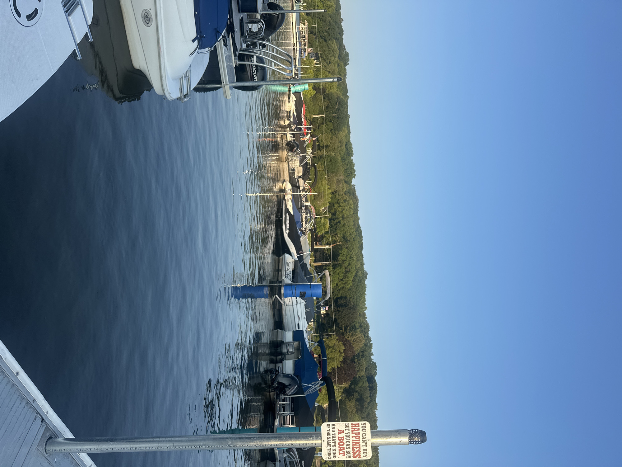 The image depicts a marina on a clear day with a bright blue sky. Several boats are docked on the water, with a focus on a row of moored vessels in the middle ground. The foreground shows a portion of a dock with a white boat and a metal railing. A sign on a pole reads, "YOU CAN'T BUY HAPPINESS BUT YOU CAN BUY A BOAT AND THAT'S KIND OF THE SAME THING." The background features a tree-lined shore with a few buildings visible amongst the foliage. The water is calm with gentle ripples reflecting the sky and the boats. The overall scene suggests a recreational area, likely a lake or river. Based on visual cues and the explicit mention of Algonquin, the location is a marina in Algonquin, United States of America. There are no people visible in the image.