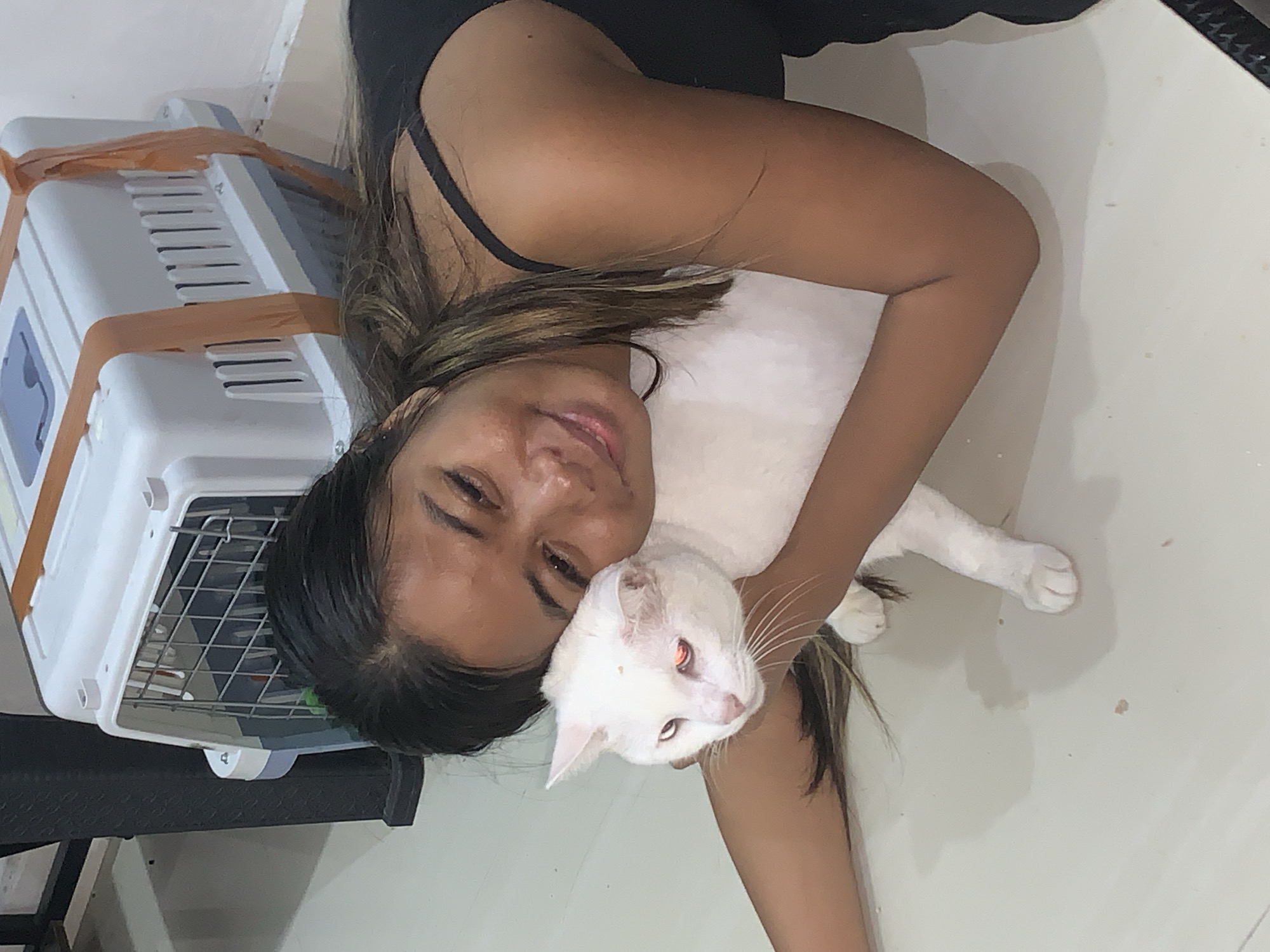 A person lies on their back, holding a white cat. The person has long, dark hair with lighter streaks, and is wearing a black tank top. They are smiling and looking at the camera. The cat is also looking towards the camera, with bright orange eyes. The cat is being held close to the person's face, with one of its paws resting on the person's chest. In the background, a white and gray pet carrier is visible, secured with an orange strap. The floor appears to be white tile. The lighting suggests it is daytime, possibly indoors. The overall mood is affectionate and playful.