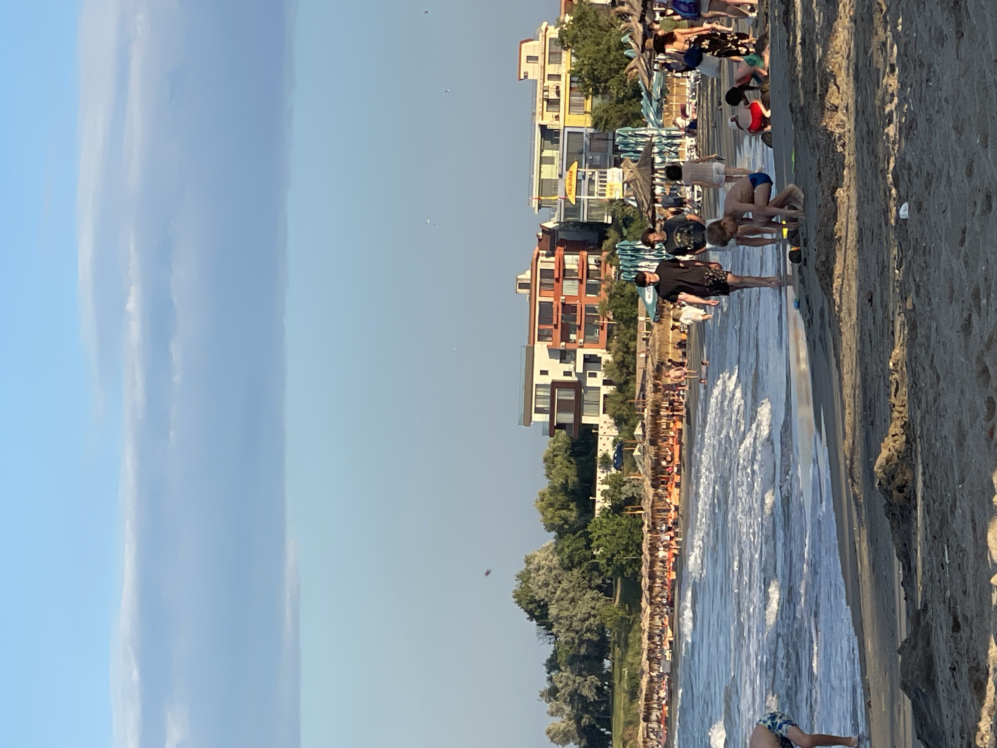 A wide-angle, eye-level, vertical shot captures a bustling beach scene on a bright day. The sky is a clear, pale blue with a thin, horizontal band of white clouds across the upper portion. The background features a row of multi-story buildings, with varying architectural styles and bright colors, including orange, yellow, and white. These buildings are situated behind a line of straw umbrellas and beach chairs, which are set up on a sandy area. Lush green trees and bushes are visible behind the buildings.
In the mid-ground, the sea is a vibrant blue-green with moderate waves lapping onto the shore. The wet sand of the beach is visible at the water's edge. People are scattered across the sand, with some walking and others interacting with the environment. In the foreground, several children are engaged in activities on the sand. One young boy, wearing blue swim trunks, is crouched down, appearing to be building or playing with sand. Nearby, another child is also playing in the sand. A group of adults and teenagers are walking along the shoreline.
The overall impression is a lively beach resort environment with clear weather.